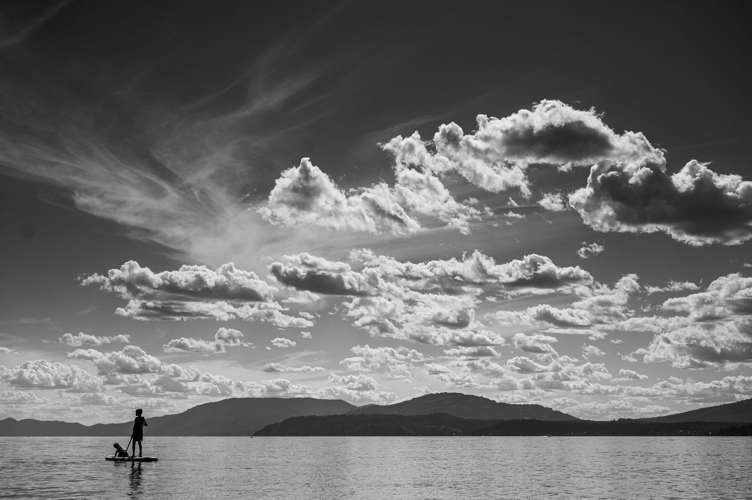 Person paddleboarding on calm lake with dramatic cloudy sky and mountain silhouette in black and white.