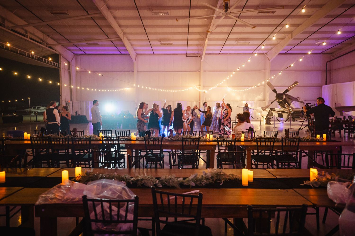 Elegant wedding reception in a warmly lit venue with string lights, candles, and guests dancing in the background.