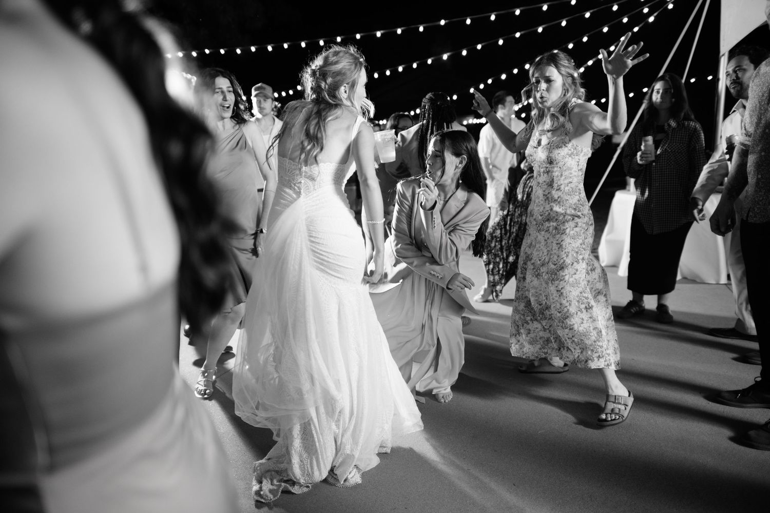 Black and white photo of wedding guests dancing under twinkling string lights at an outdoor reception.