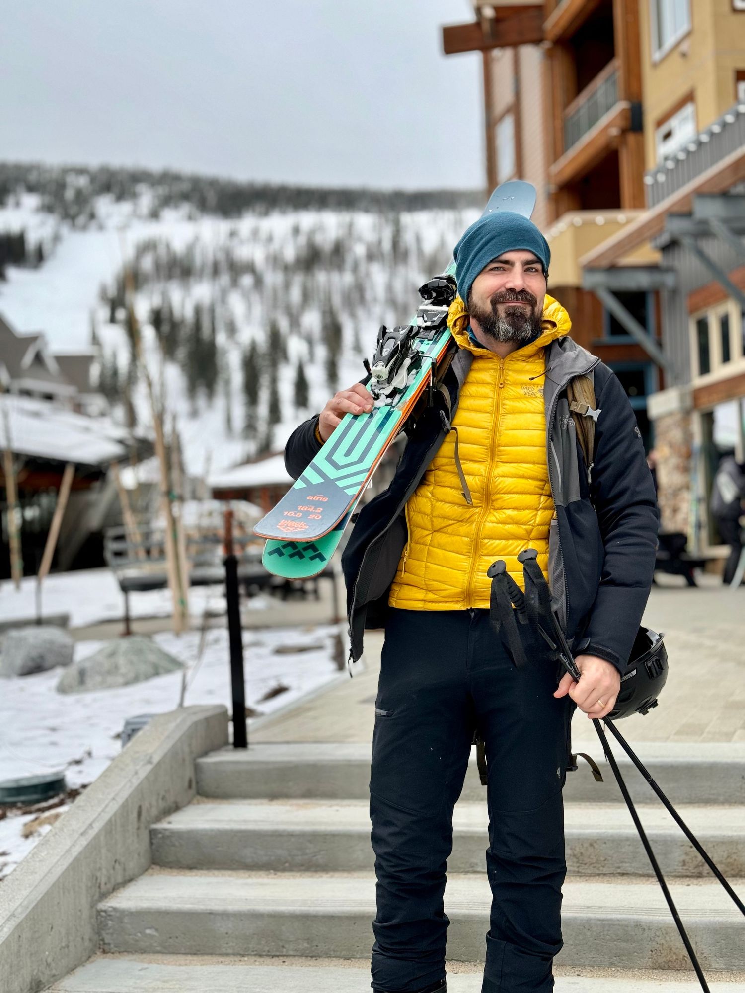 Skier in yellow puffy jacket and blue hat carrying equipment on snowy resort steps.