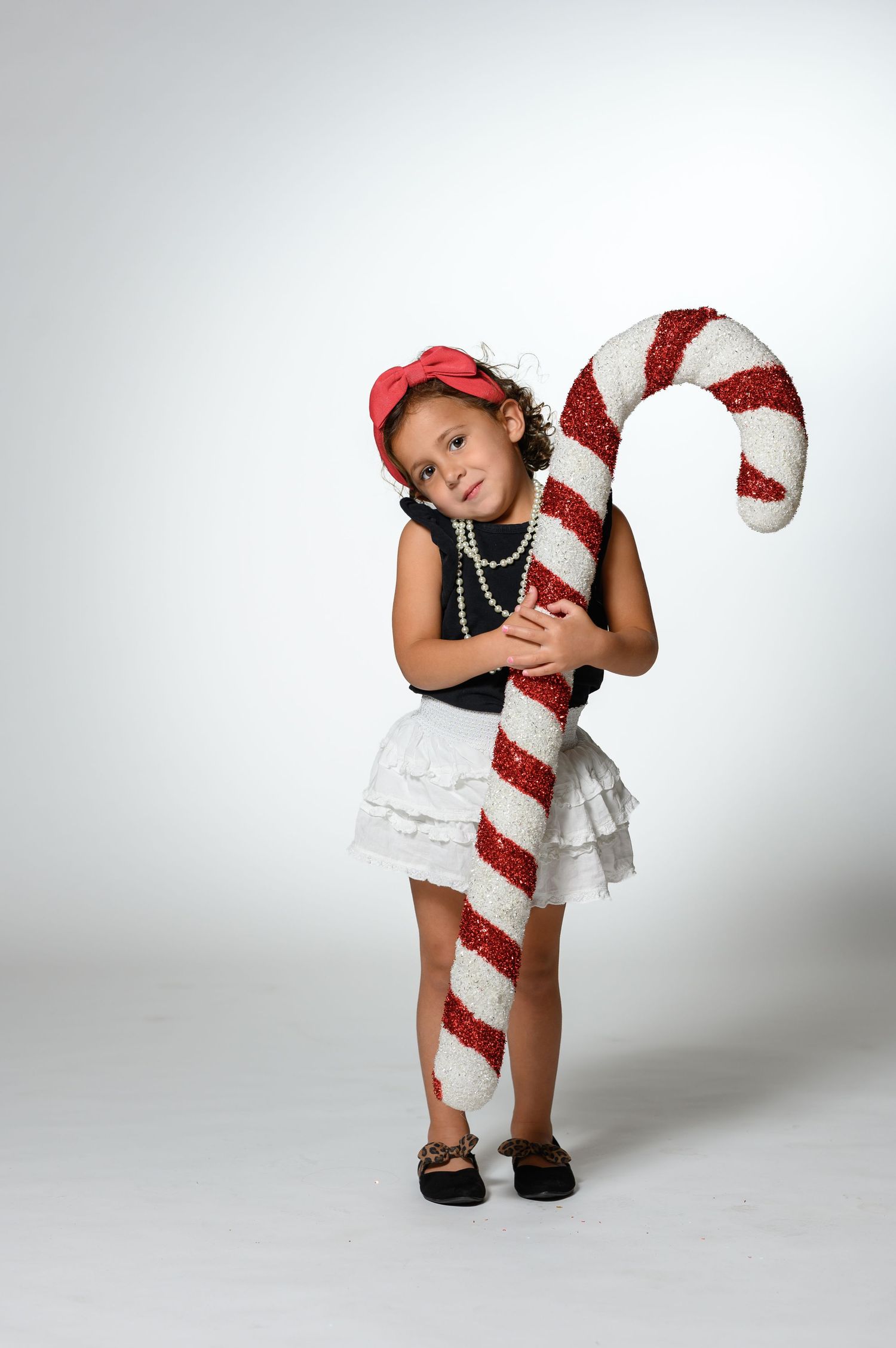 Young girl in white skirt and red bow hugging a large decorative red and white candy cane prop.