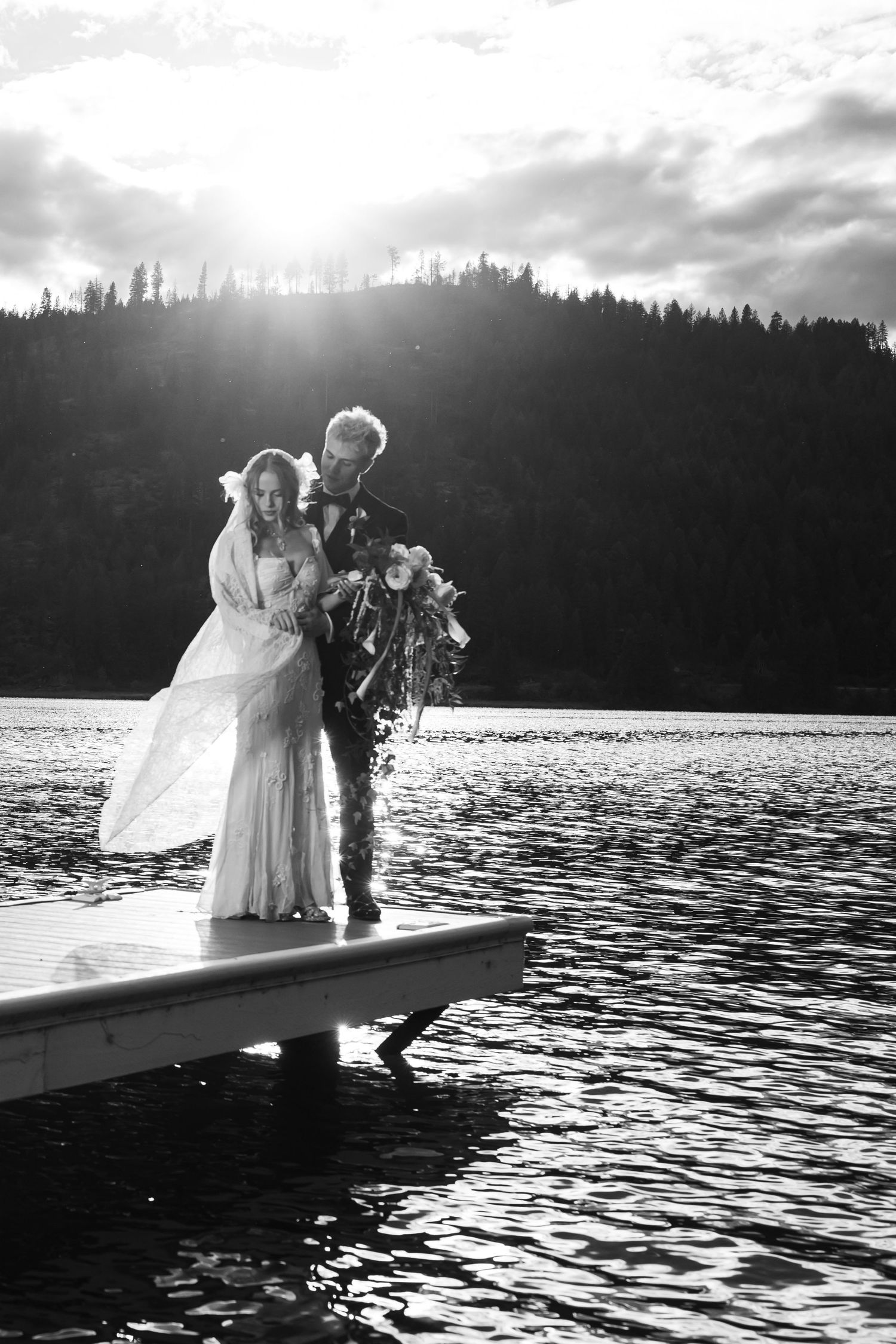 Bride and groom stand on a lake dock at sunset in a romantic black and white wedding portrait.