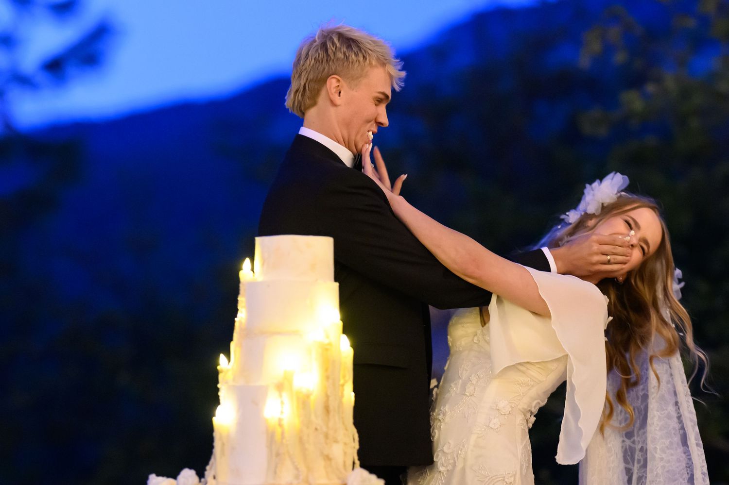 Groom playfully covers bride's face at outdoor candlelit wedding cake cutting ceremony at dusk.