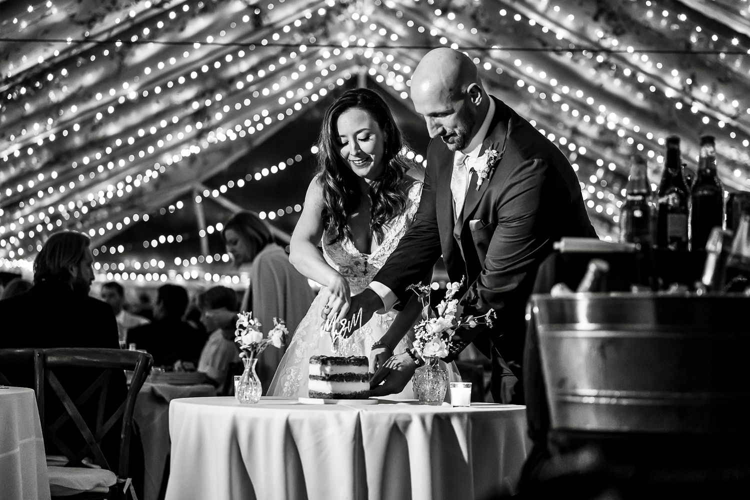 A black and white photo of a wedding couple cutting their cake together beneath sparkling fairy lights.
