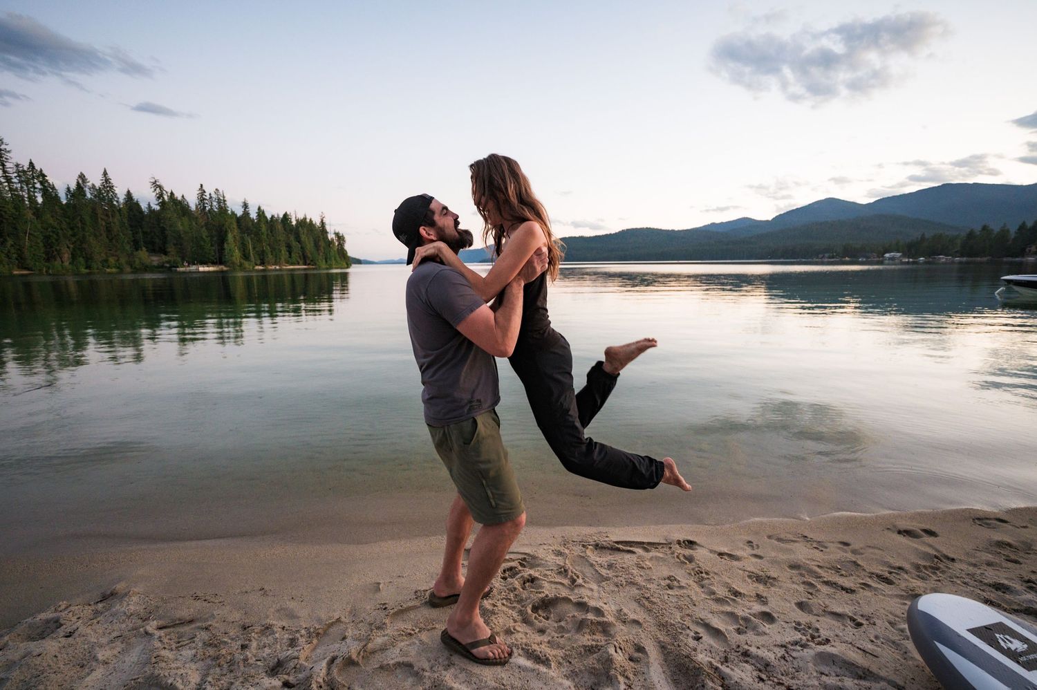 A playful couple embraces at sunset on a sandy beach with mountains and pine forest reflected in the calm lake water.