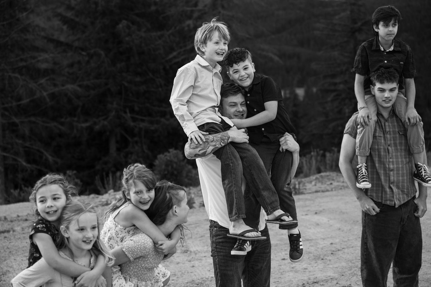 Black and white family photo of adults and children playing outdoors near pine trees on sandy ground.