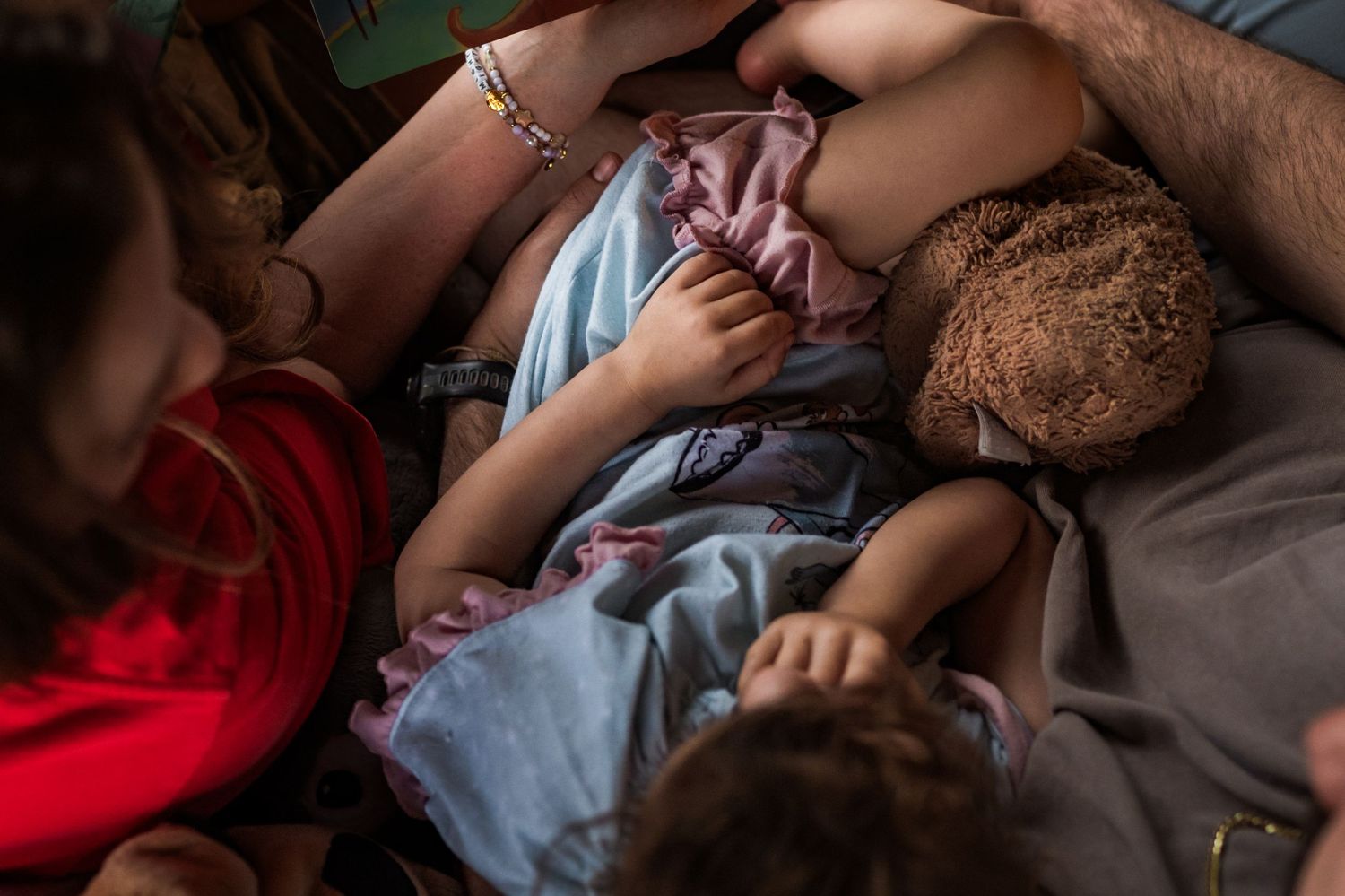 Mother breastfeeding a toddler holding a teddy bear while relaxing indoors.