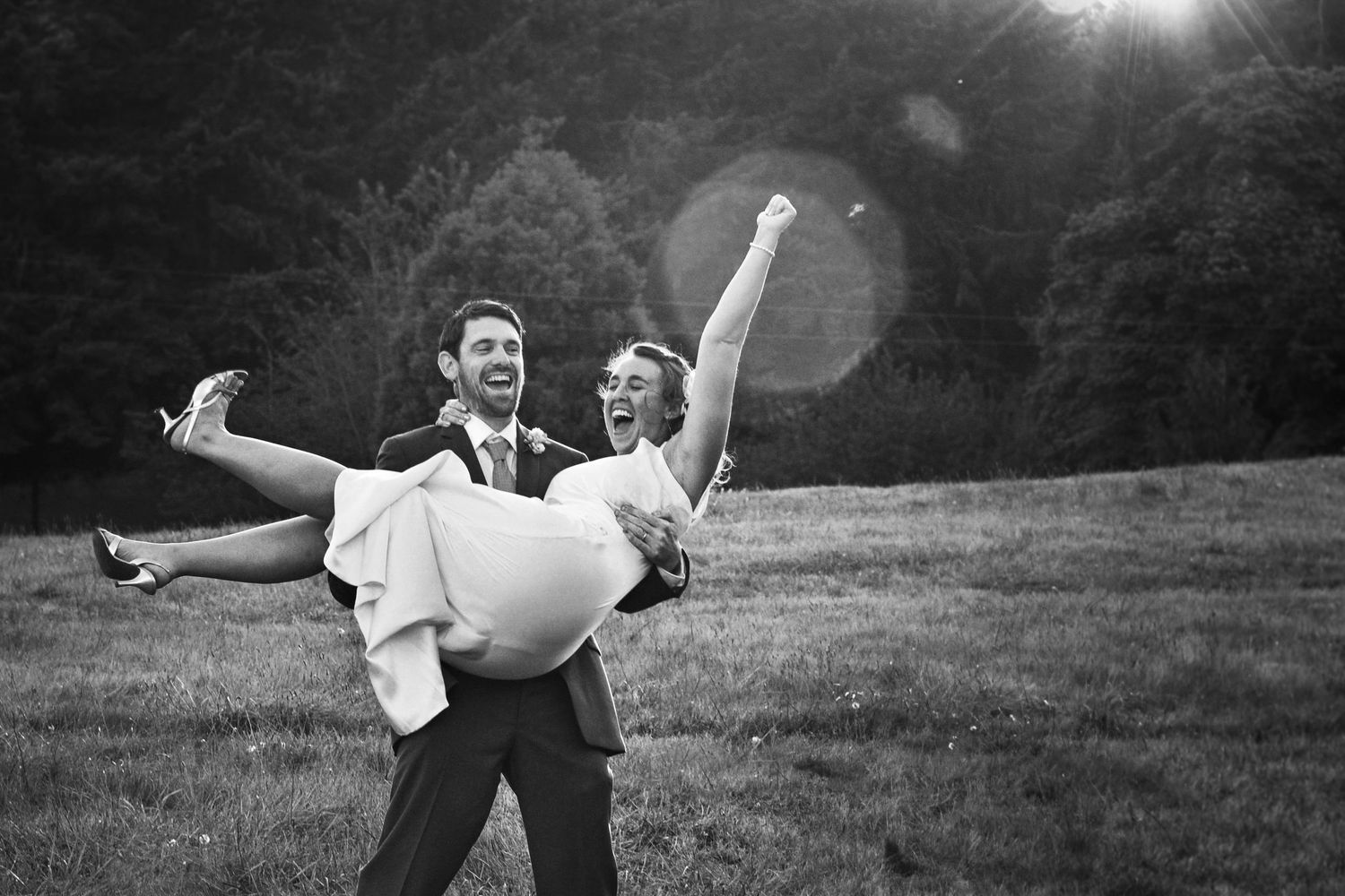 Joyful groom carries laughing bride in wedding attire outdoors in black and white photo with sun flare.