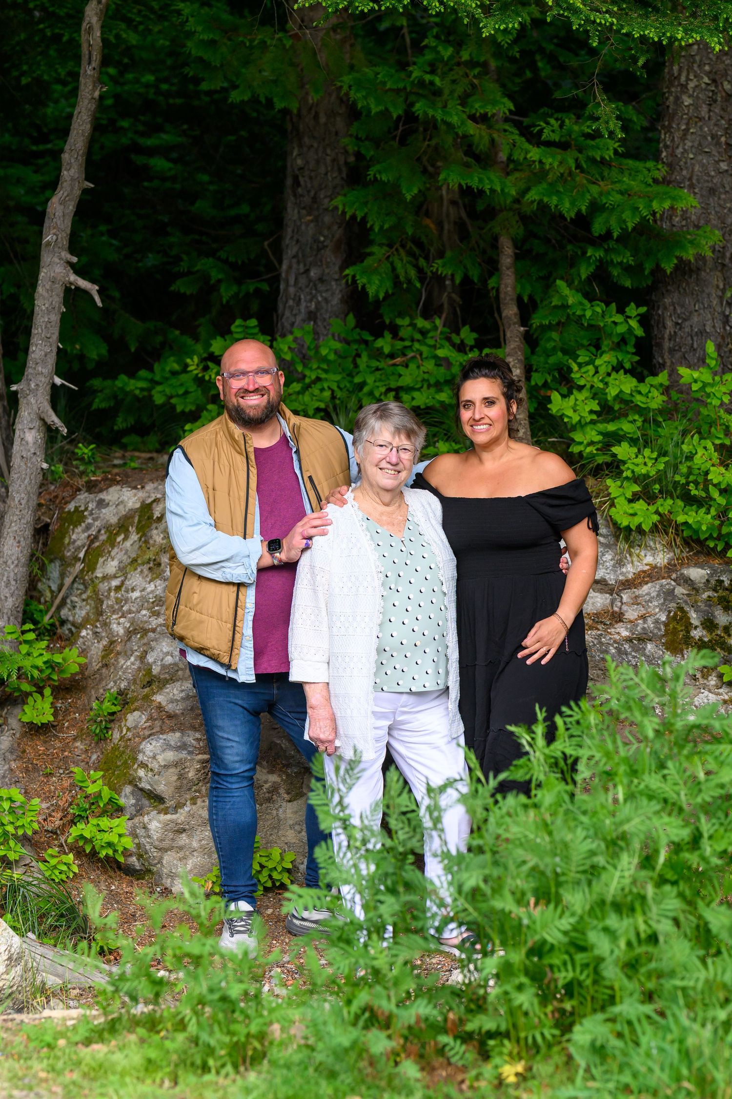 A bearded man, elderly woman, and brunette woman smile together outdoors in a wooded setting.