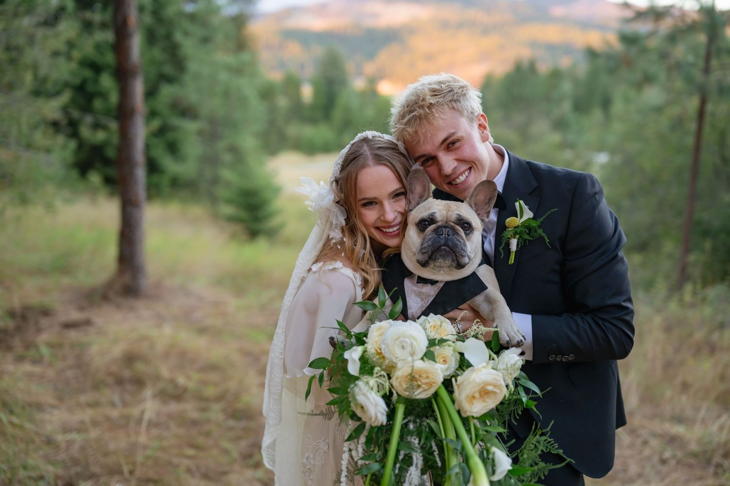 Smiling bride and groom in wedding attire hold a French bulldog and white floral bouquet in a forest setting.