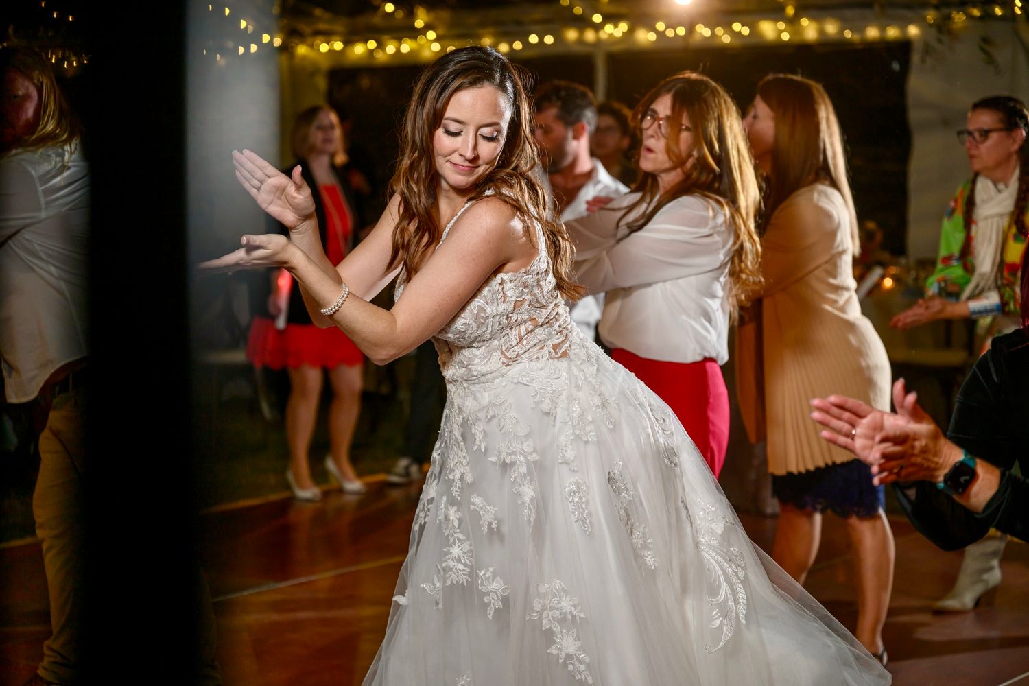 A person in a white lace wedding gown dances joyfully at a reception under twinkling string lights.