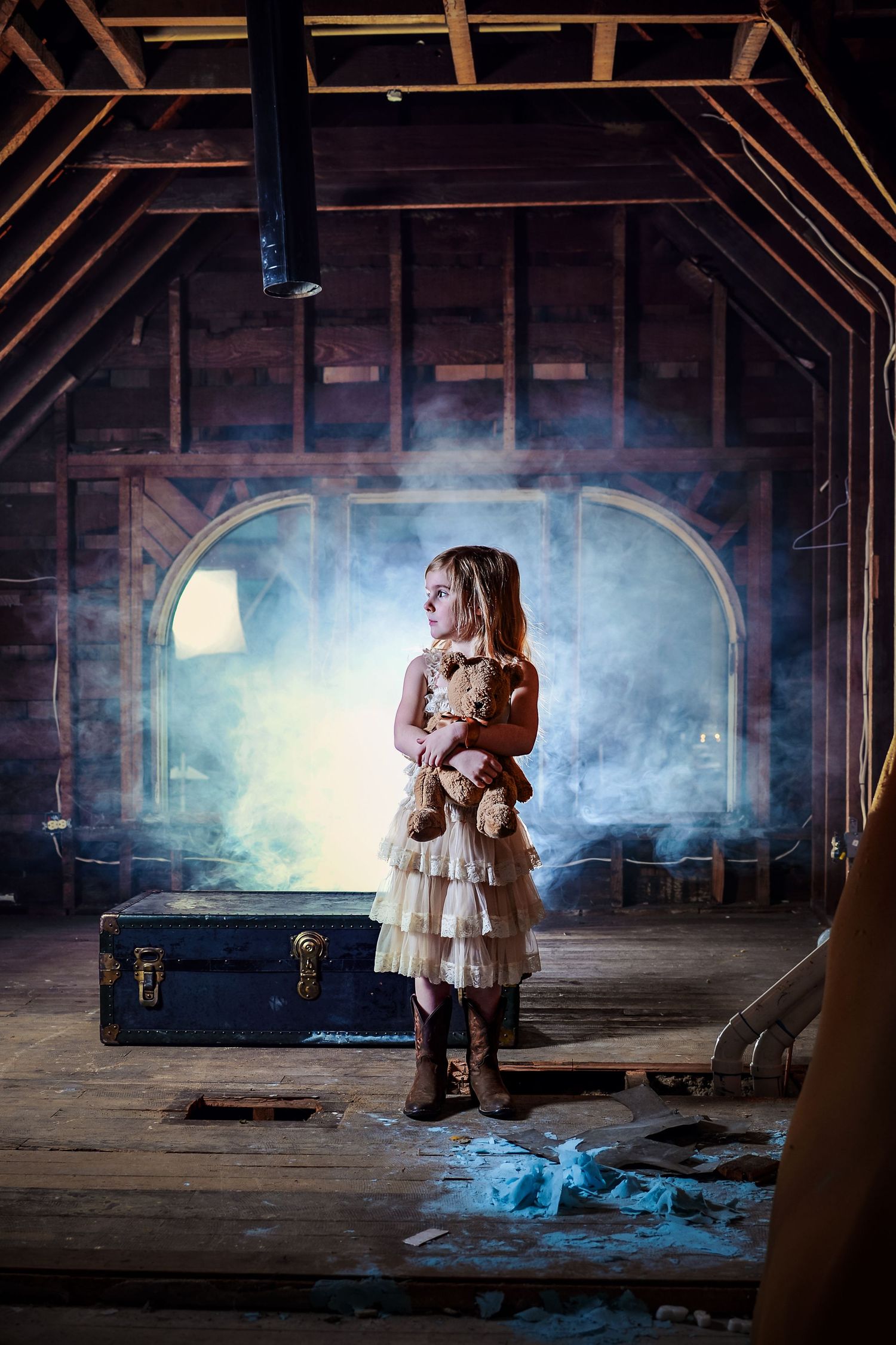 Young girl in vintage dress holding teddy bear stands in misty attic with trunk and wooden beams.
