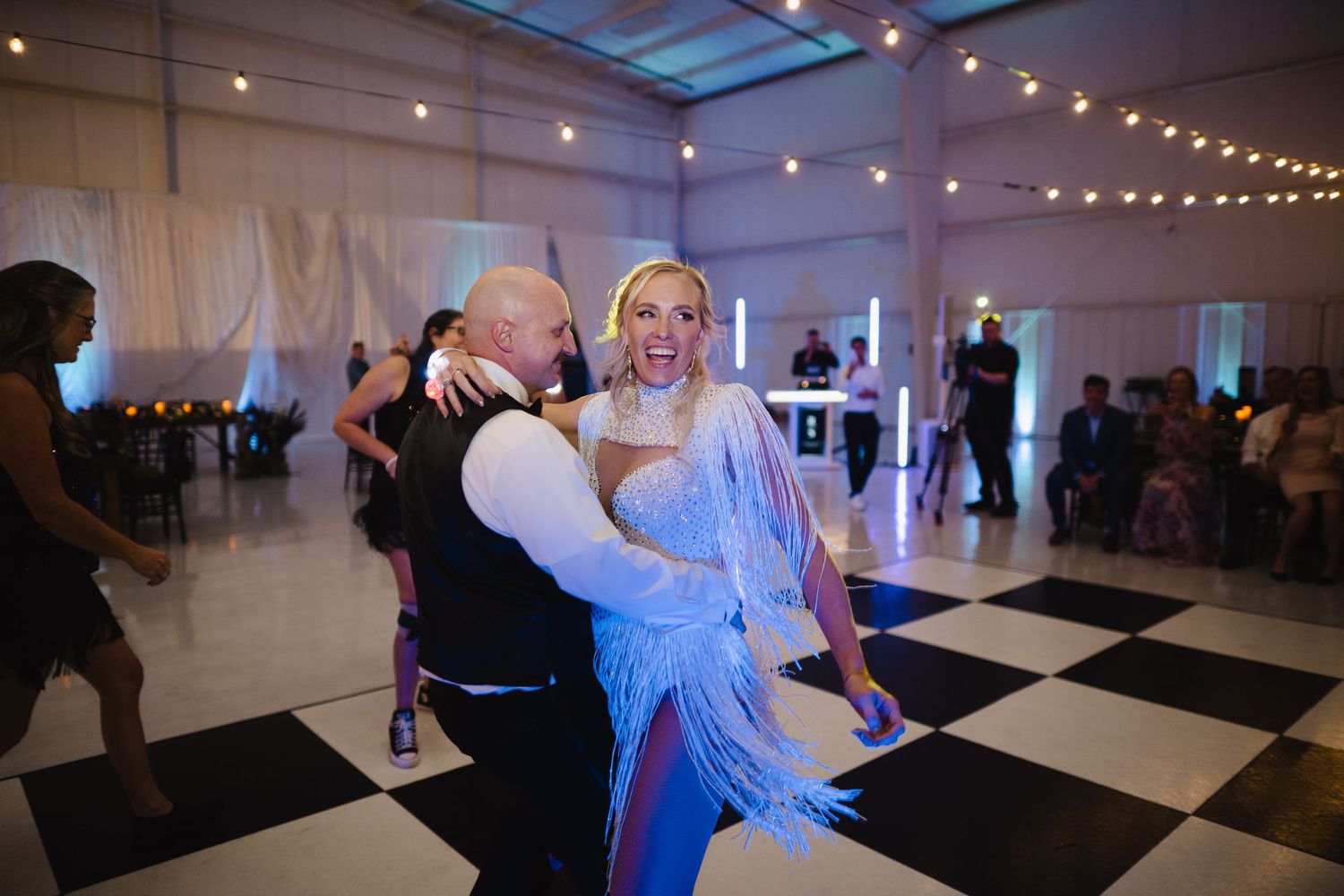 Bride in blue fringe dress laughing while being lifted by groom on black and white checkered dance floor.