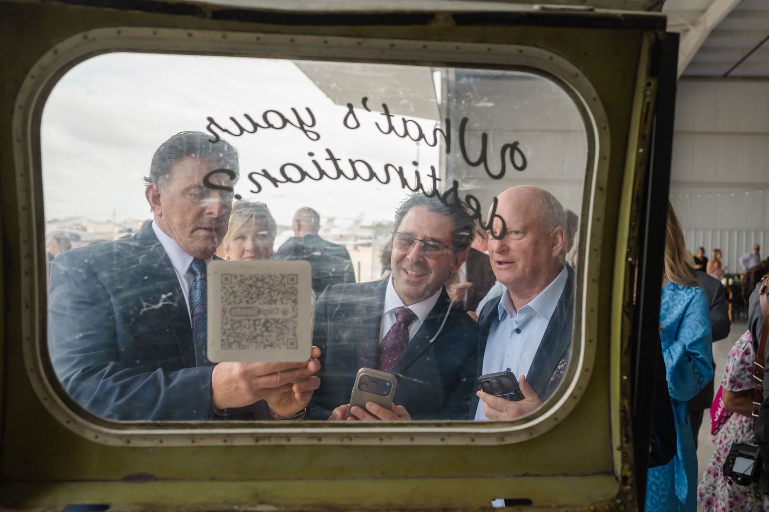 Three men smile while looking at a phone through a military vehicle window at an airshow.
