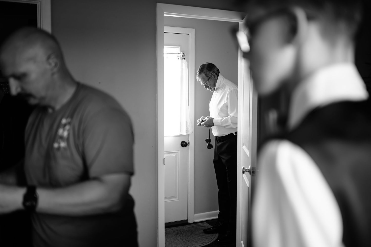 Black and white wedding photo of groomsmen preparing, man adjusting cufflinks in doorway with soft natural light.