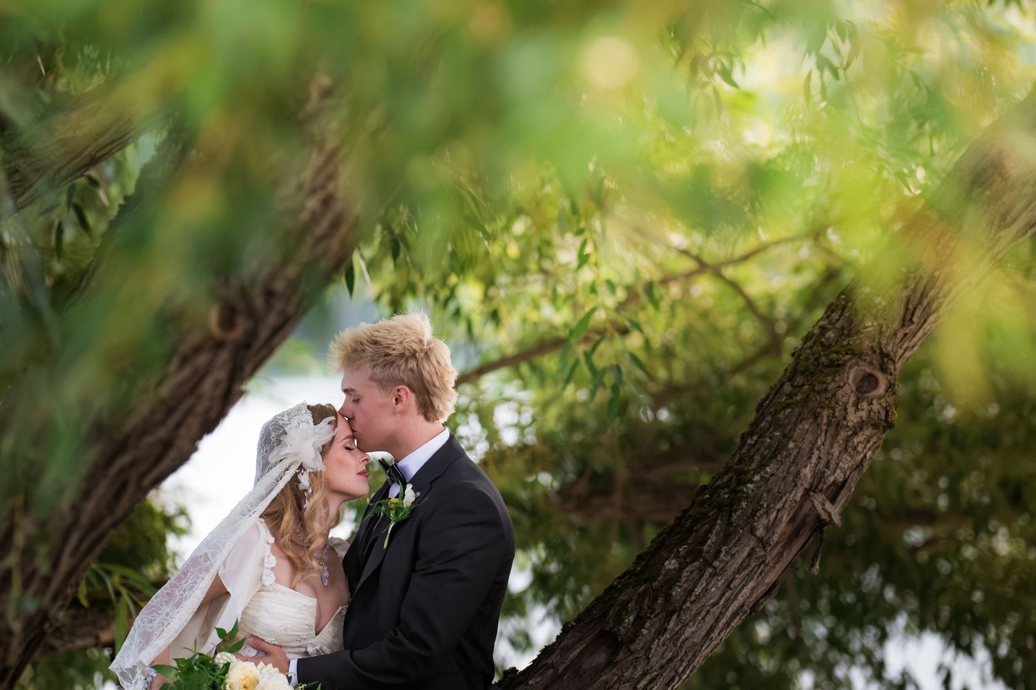 Bride and groom sharing a romantic kiss surrounded by lush green trees on their wedding day.