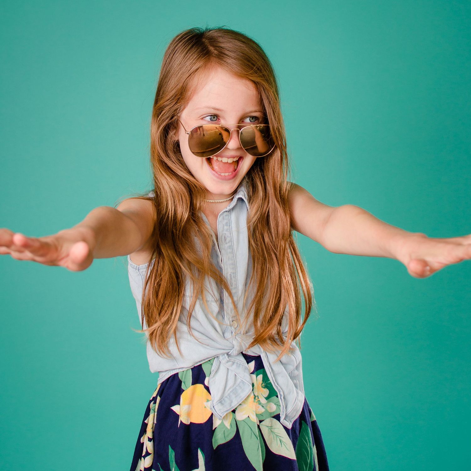 A person with long hair dances joyfully in a gray top and floral dress against a turquoise backdrop.