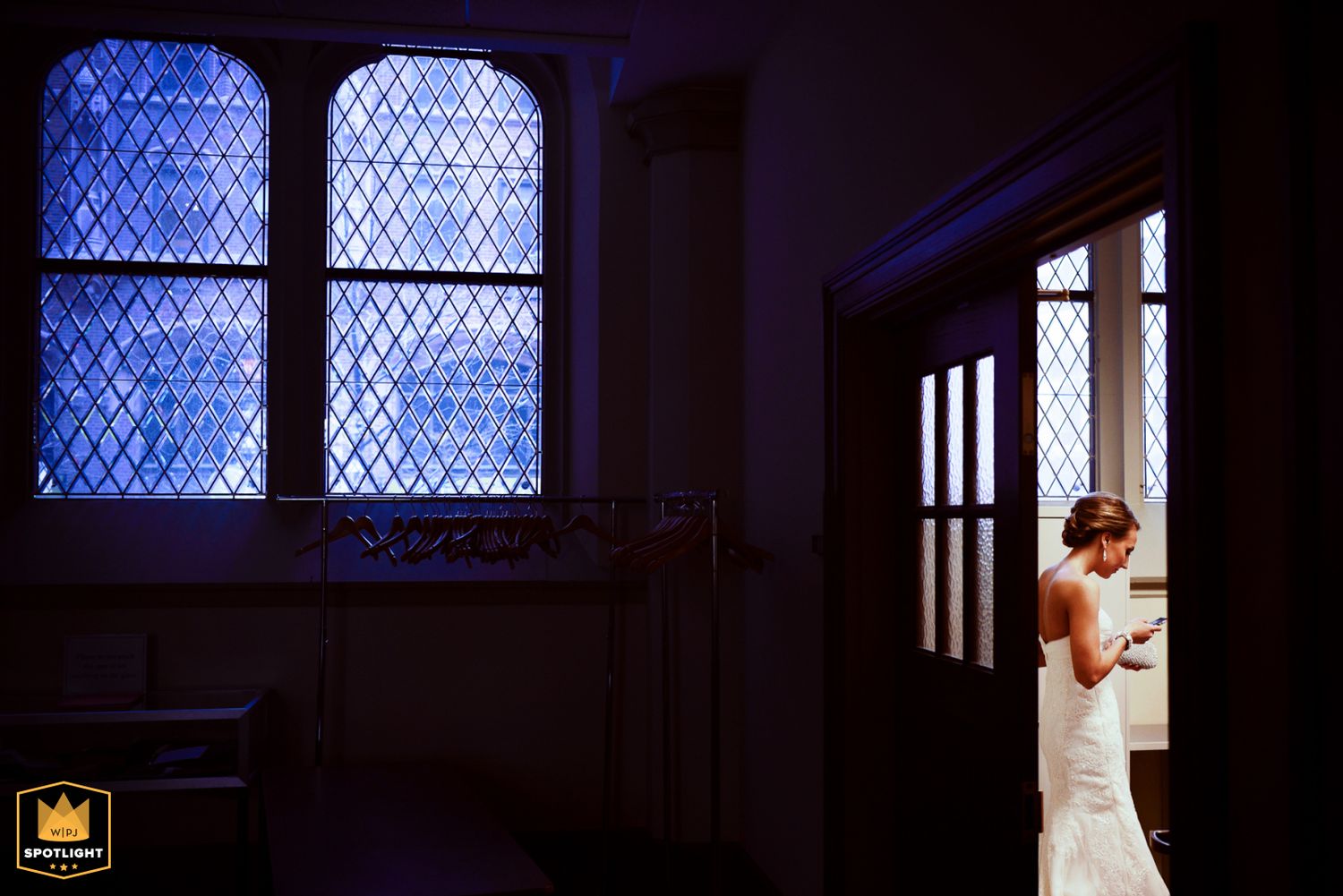 Elegant figure in a white wedding dress stands illuminated in a doorway beside blue stained glass church windows, award winning wedding pic