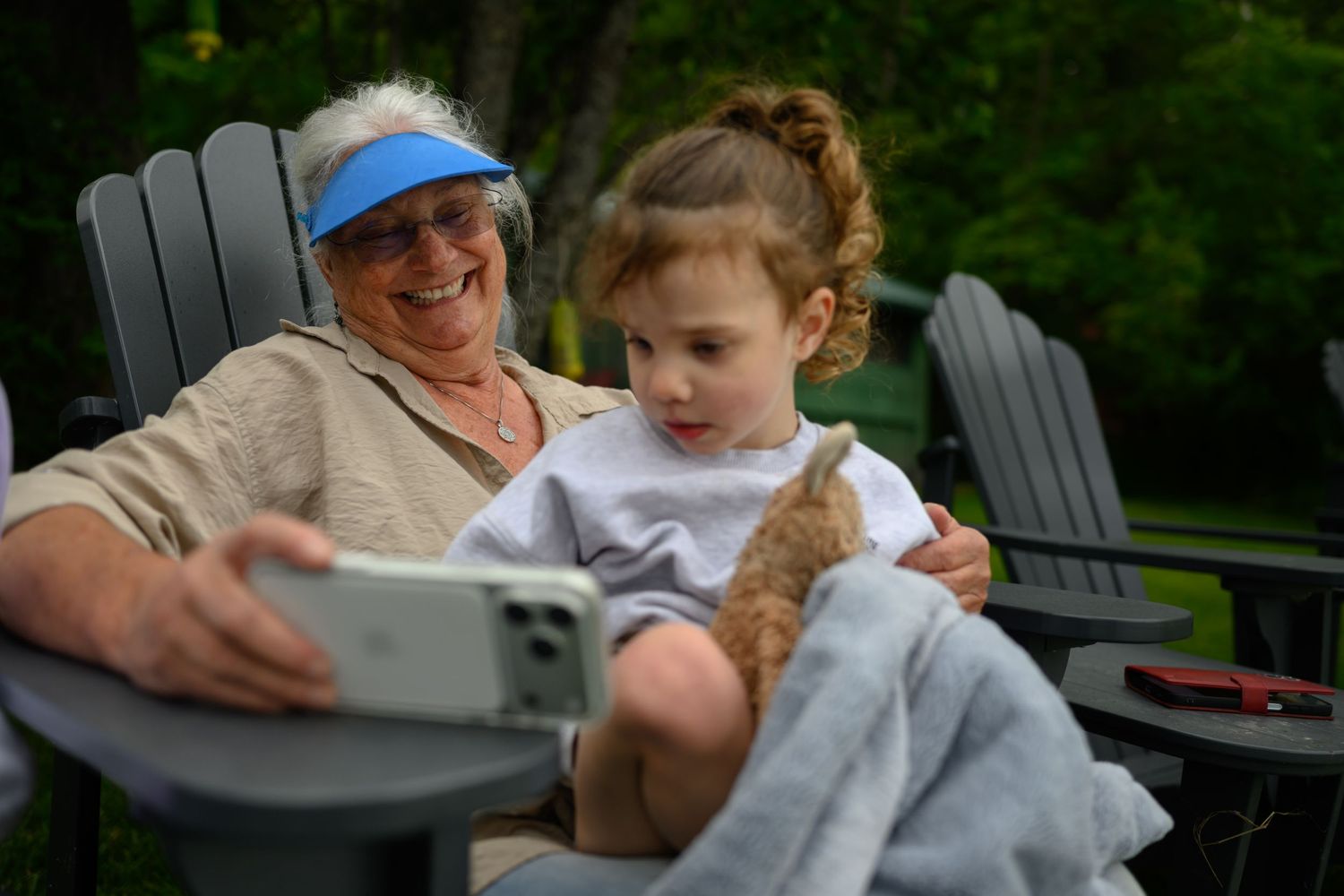Grandmother and granddaughter taking a selfie together outdoors while holding a small teddy bear.