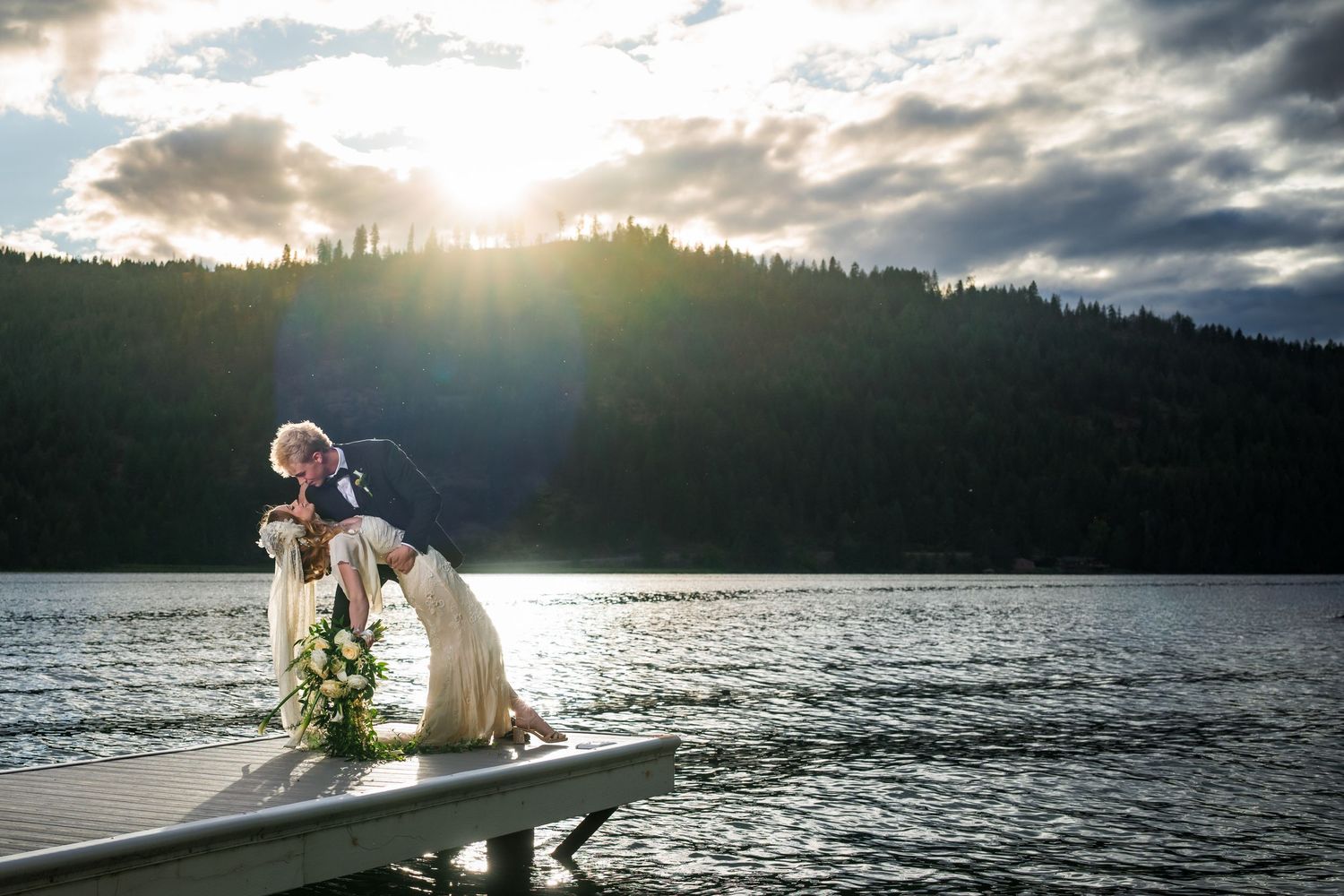 Couple sharing a romantic dip kiss on a lake dock at sunset, with lush forested hills and dramatic clouds in the background.