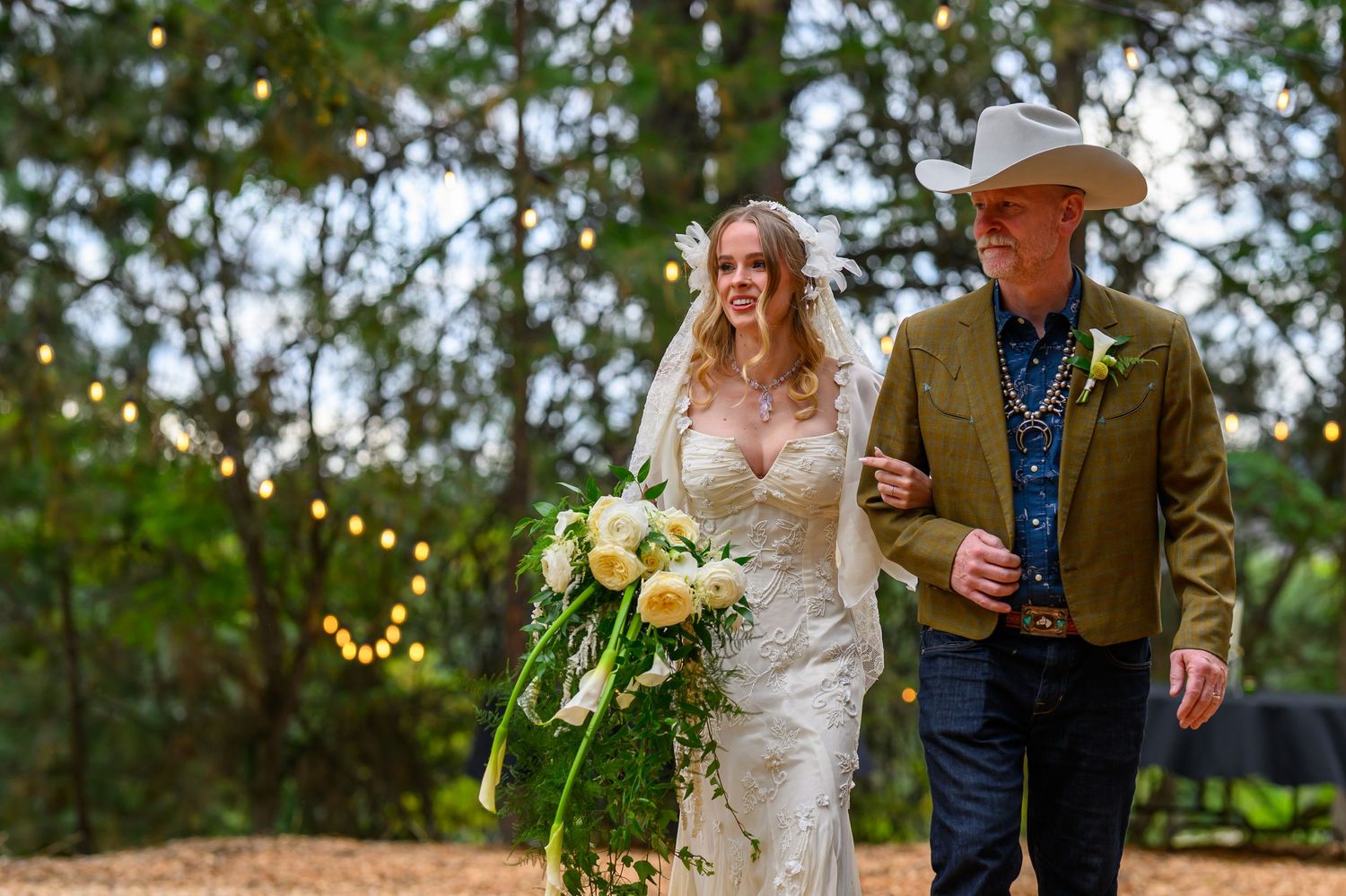 Bride in lace gown holding yellow bouquet walks with man in green jacket amid string lights at outdoor wedding.