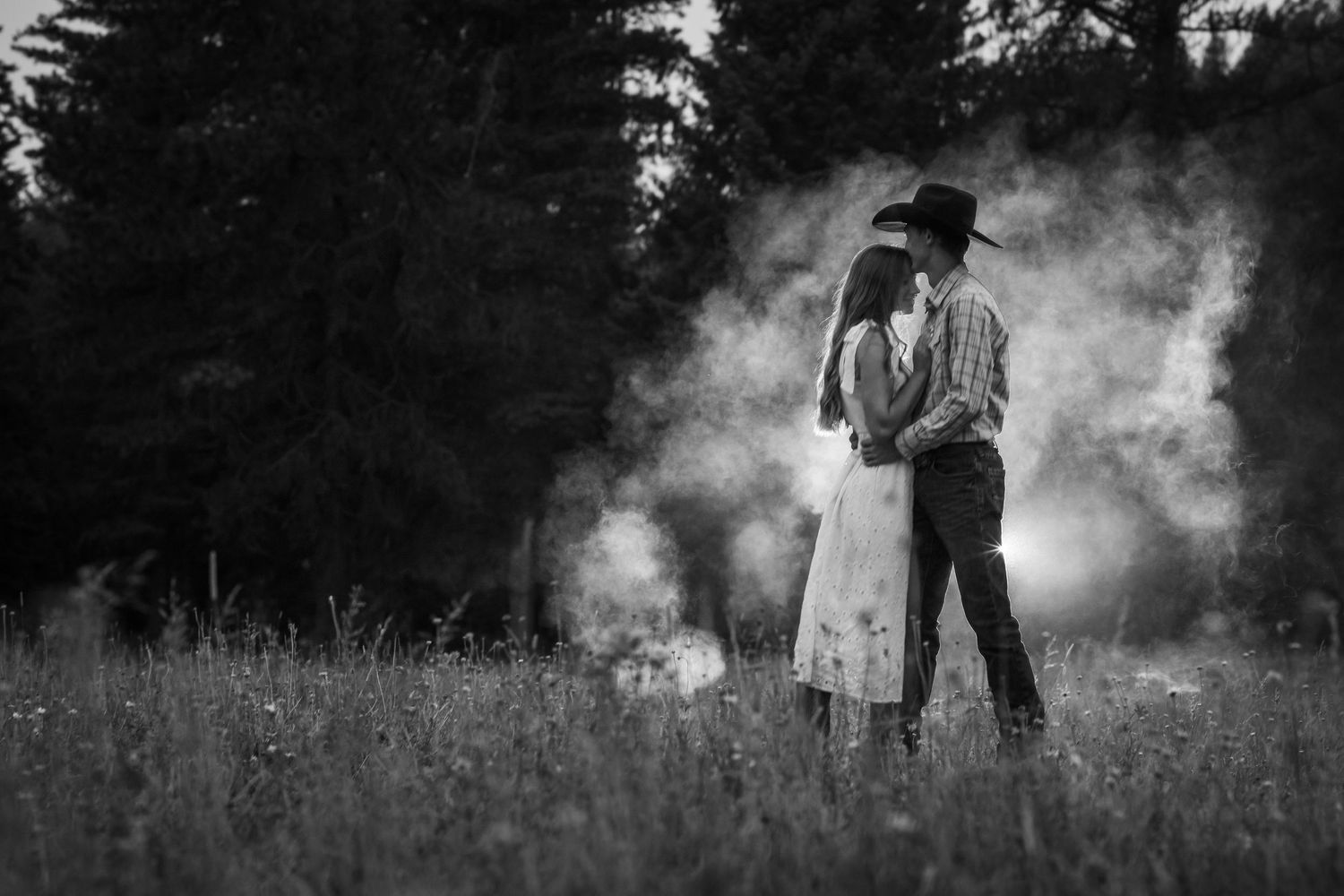 Black and white photo of a couple embracing in a misty field with trees in the background.