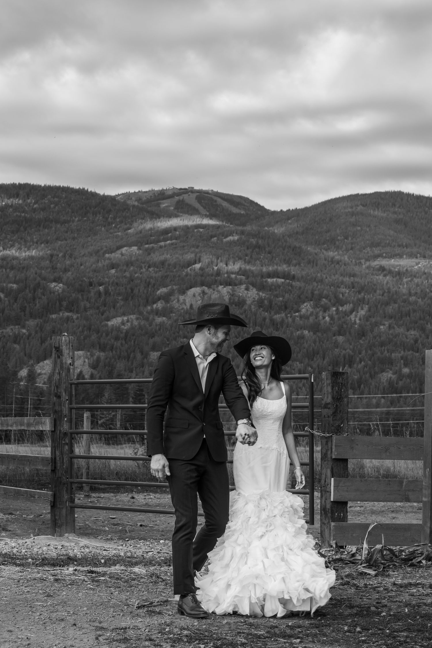 Black and white photo of a bride and groom in western hats holding hands with mountain backdrop.