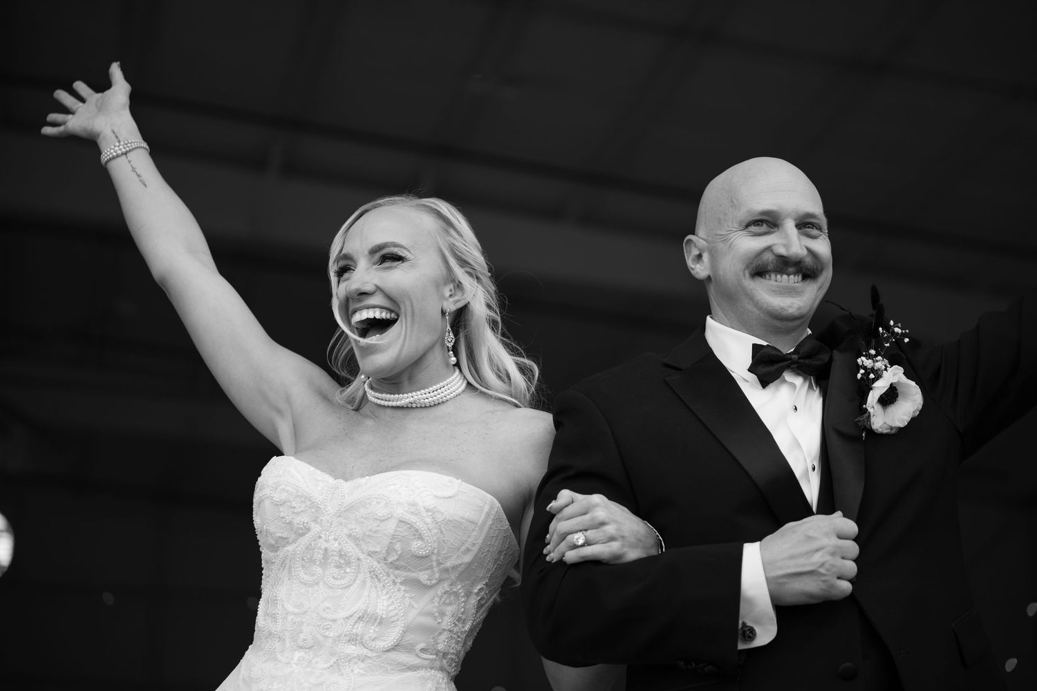 Black and white photo of a joyful bride raising her arm and laughing beside a man in a tuxedo at a wedding reception.