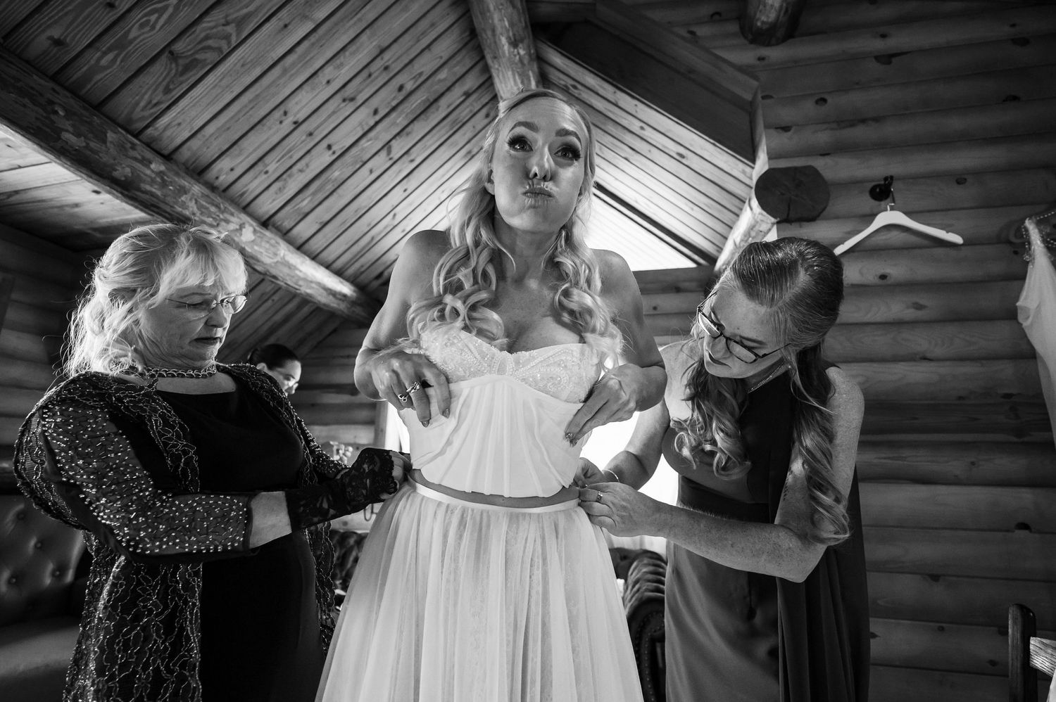 Black and white photo of a bride being helped into her wedding dress by two women in a rustic log cabin setting.