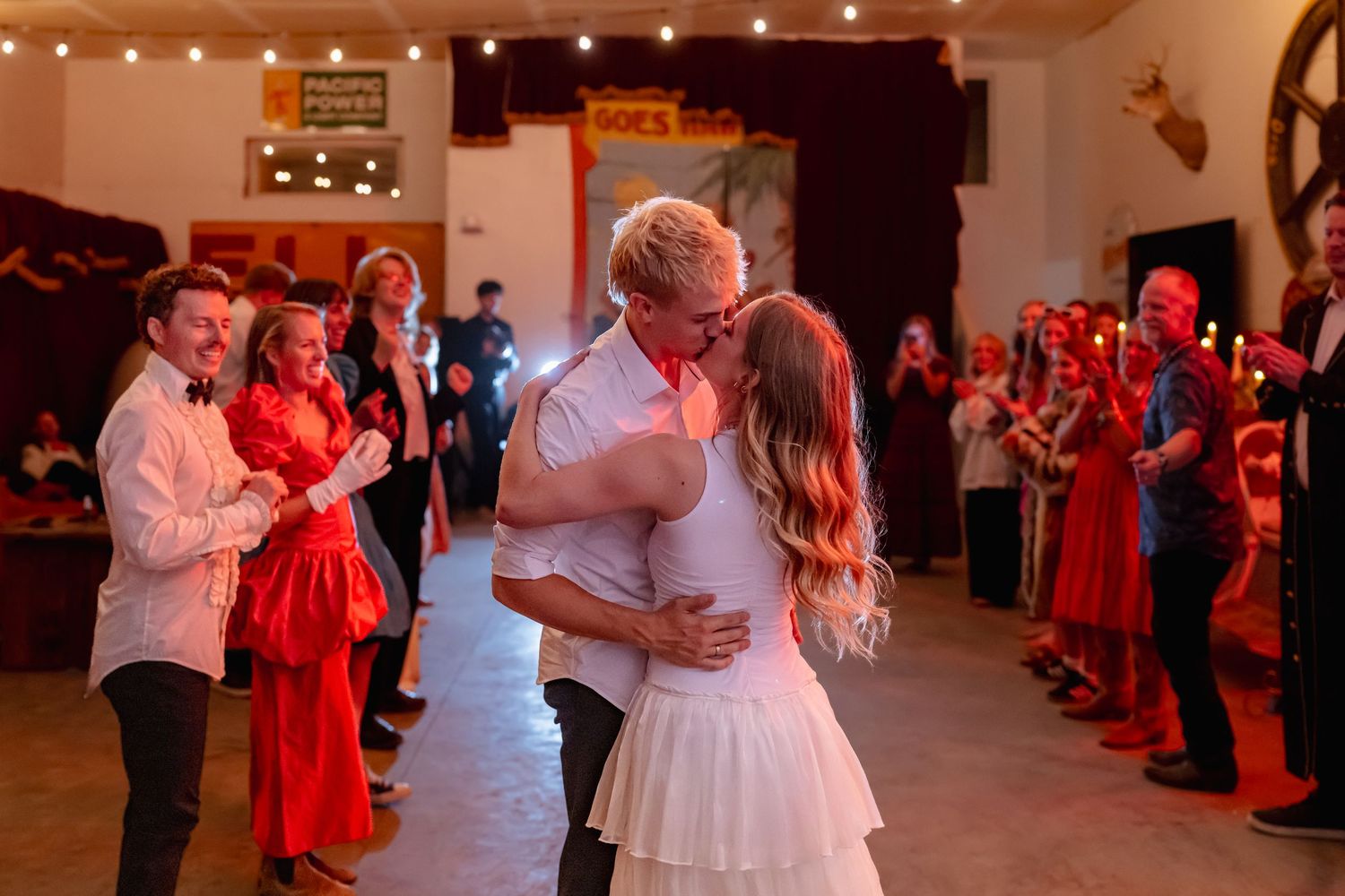 Bride and groom share first dance kiss in rustic hall surrounded by celebrating guests.