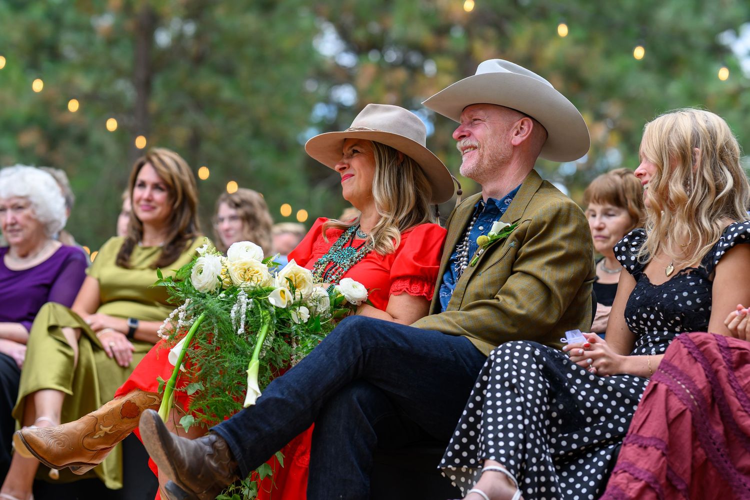 A smiling couple in cowboy hats sit together at an outdoor wedding, holding white flowers at a rustic celebration.