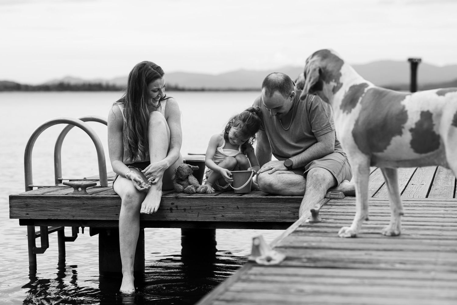Black and white photo of family with dog sitting together on a wooden lake dock.