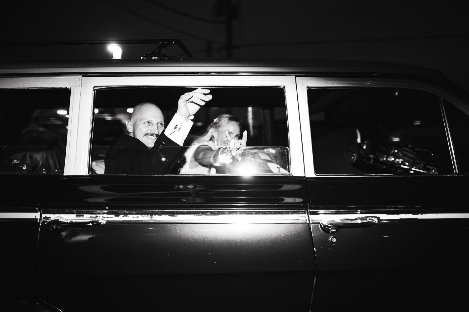 Black and white photo of two people waving from inside a classic vintage car window at night.
