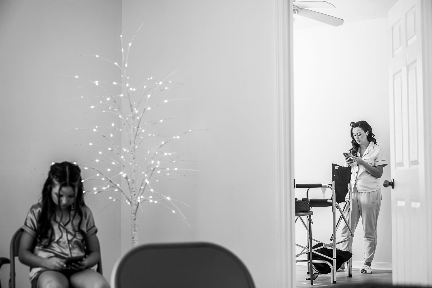 Black and white photo showing a lit up decorative tree by a doorway while someone stands in pajamas in a distant room.