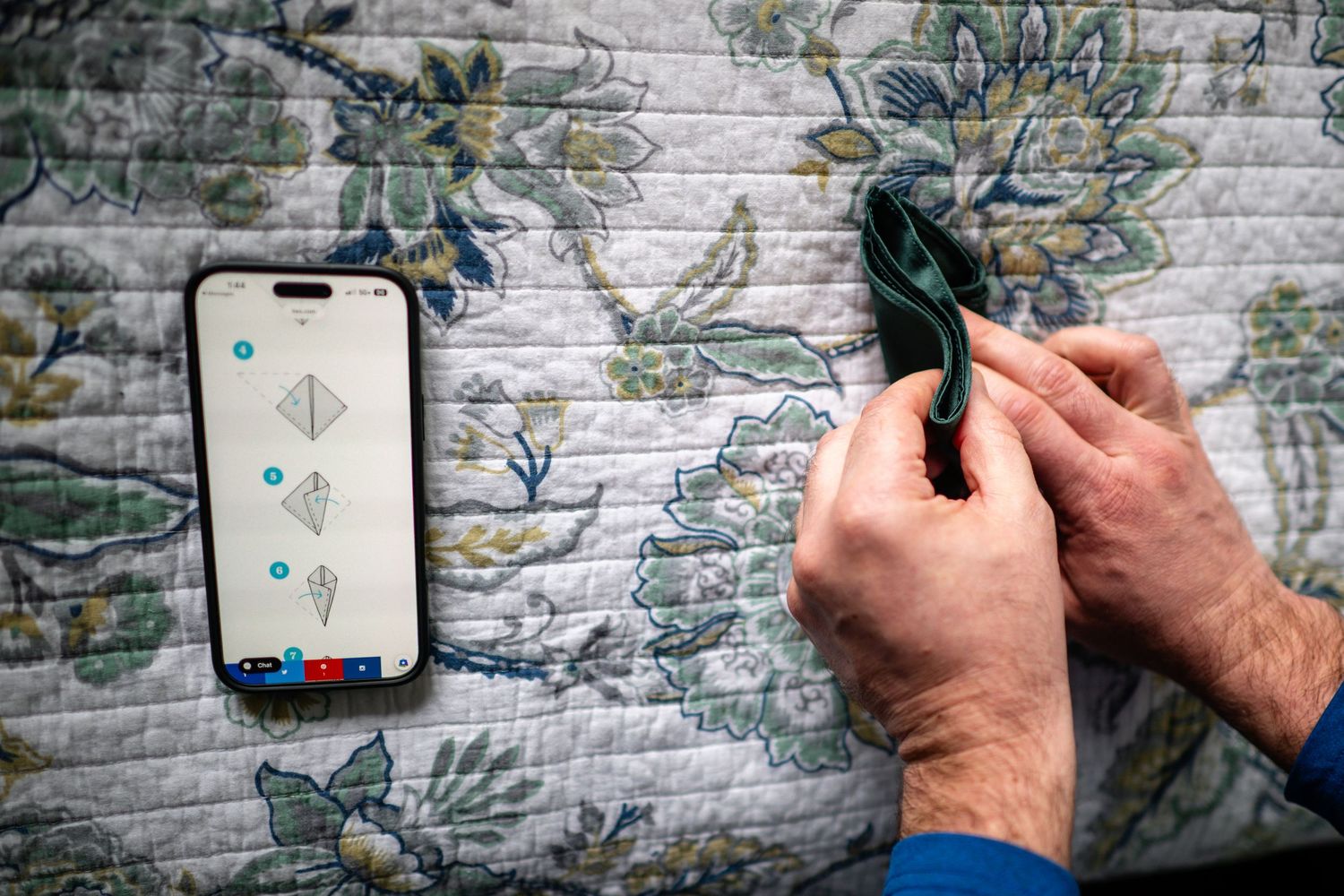 Person folding origami while following instructions on iPhone screen on floral quilt.