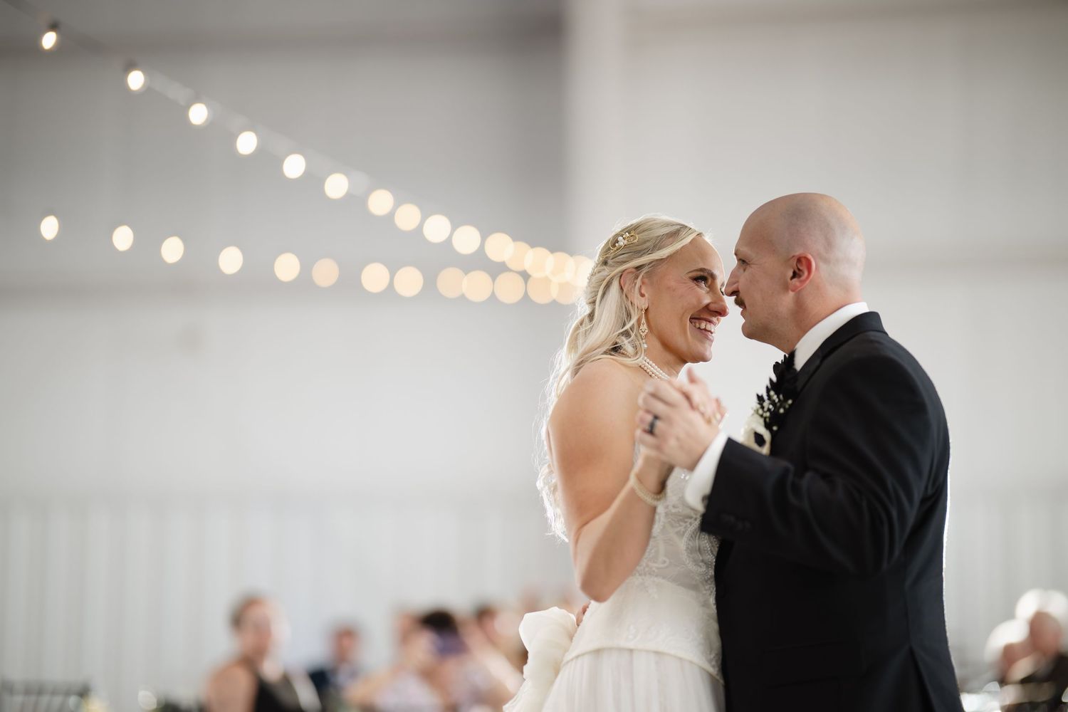 Bride and groom share their first dance, smiling joyfully under warm string lights at their wedding reception.