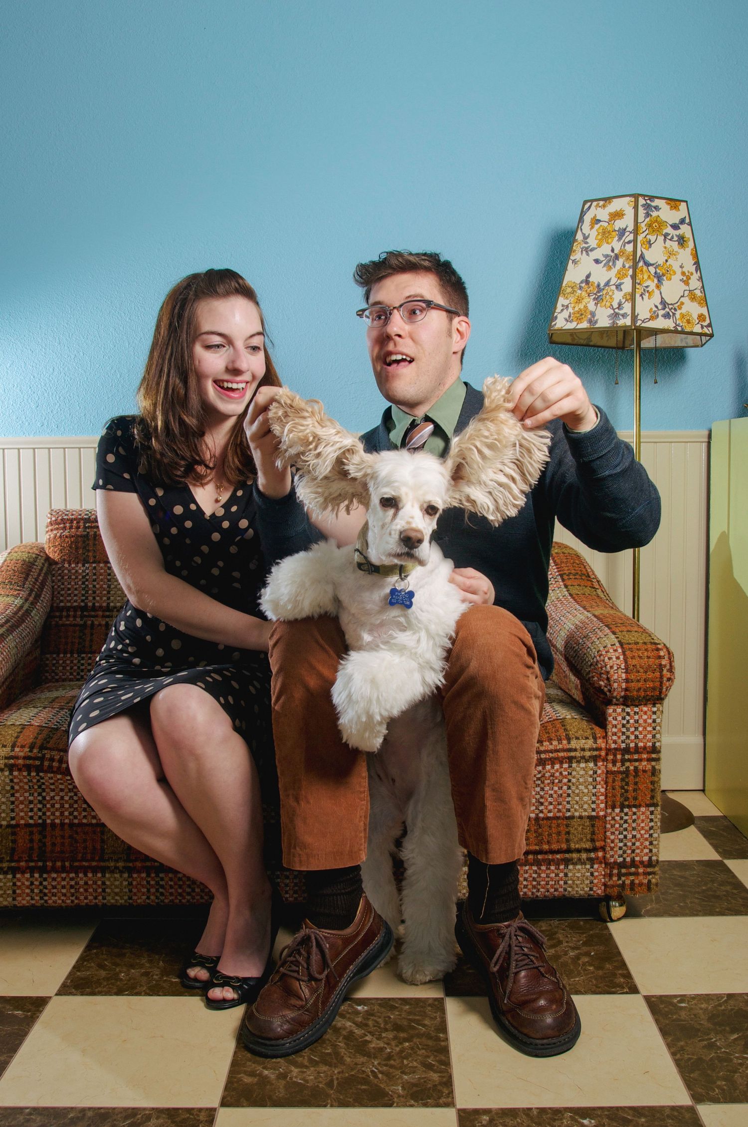 A couple sits on a plaid sofa with a white poodle, man humorously lifting the dog's ears, beside a vintage floral lamp.