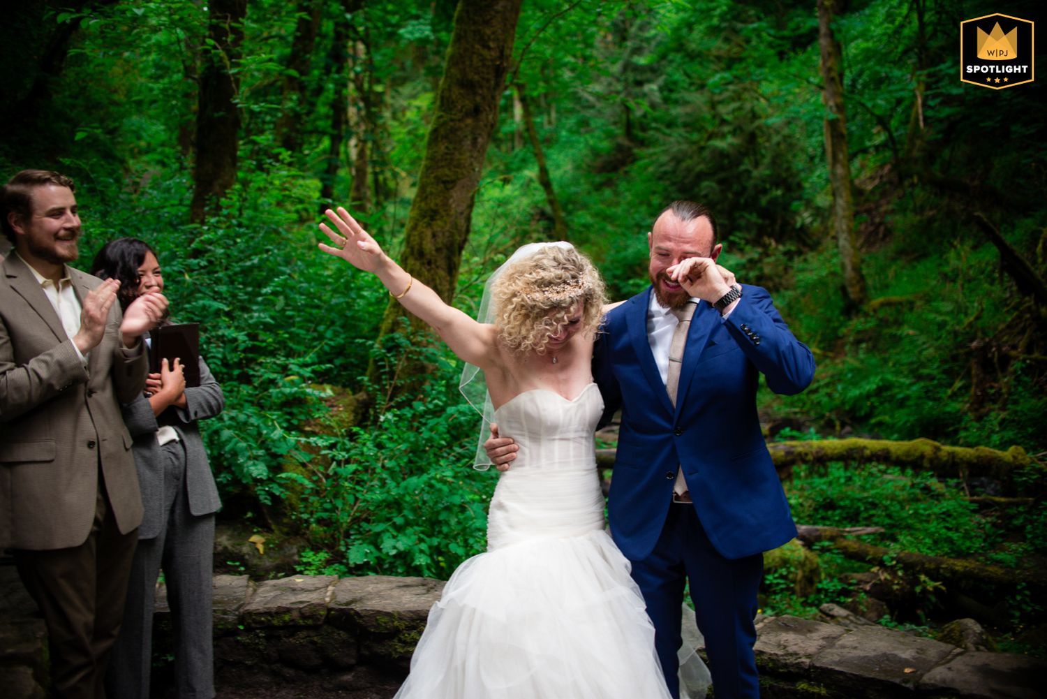 A wedding couple shares a joyful moment dancing together in a lush green forest setting.