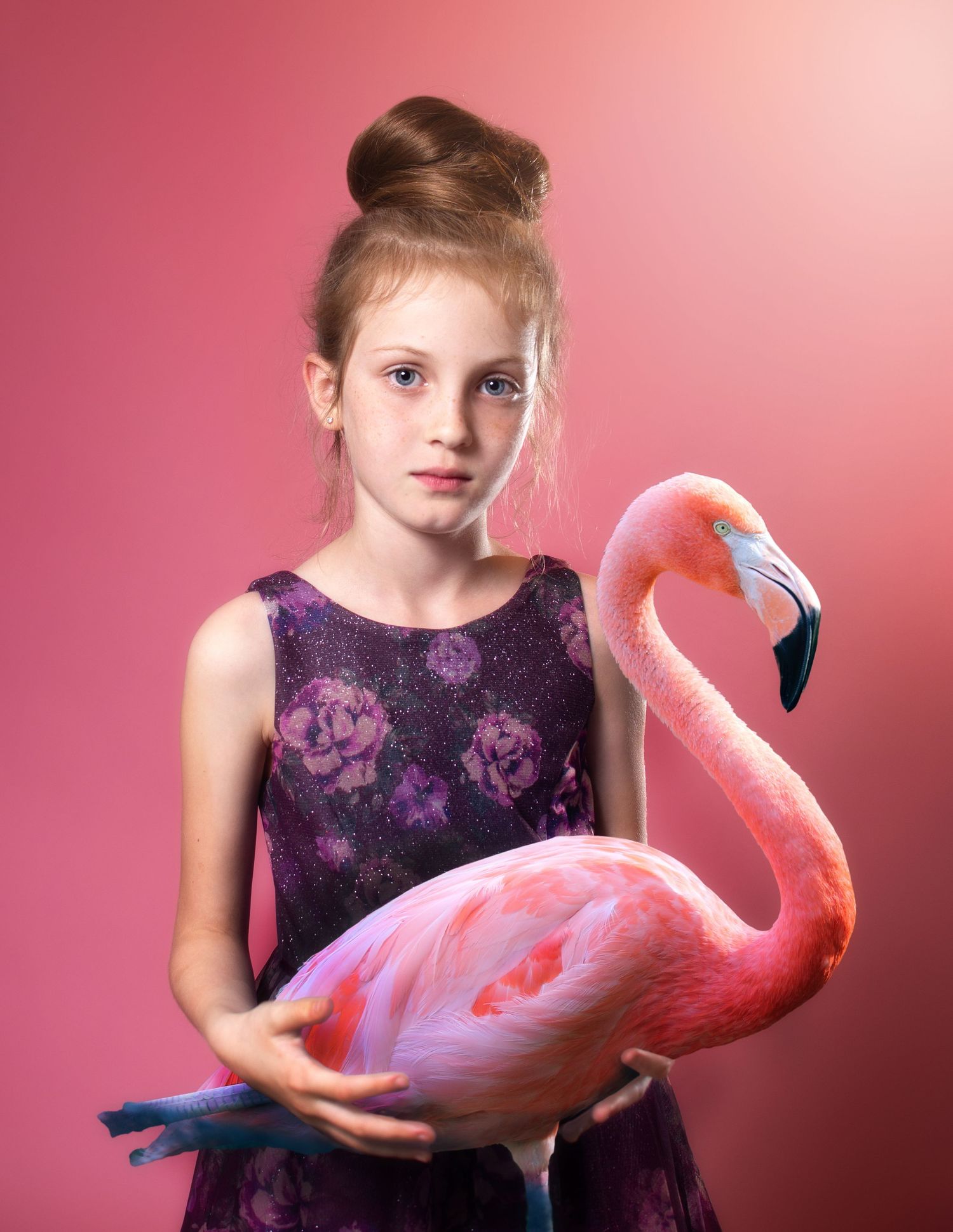 A young girl in a purple dress poses with a pink flamingo prop against a rosy background.