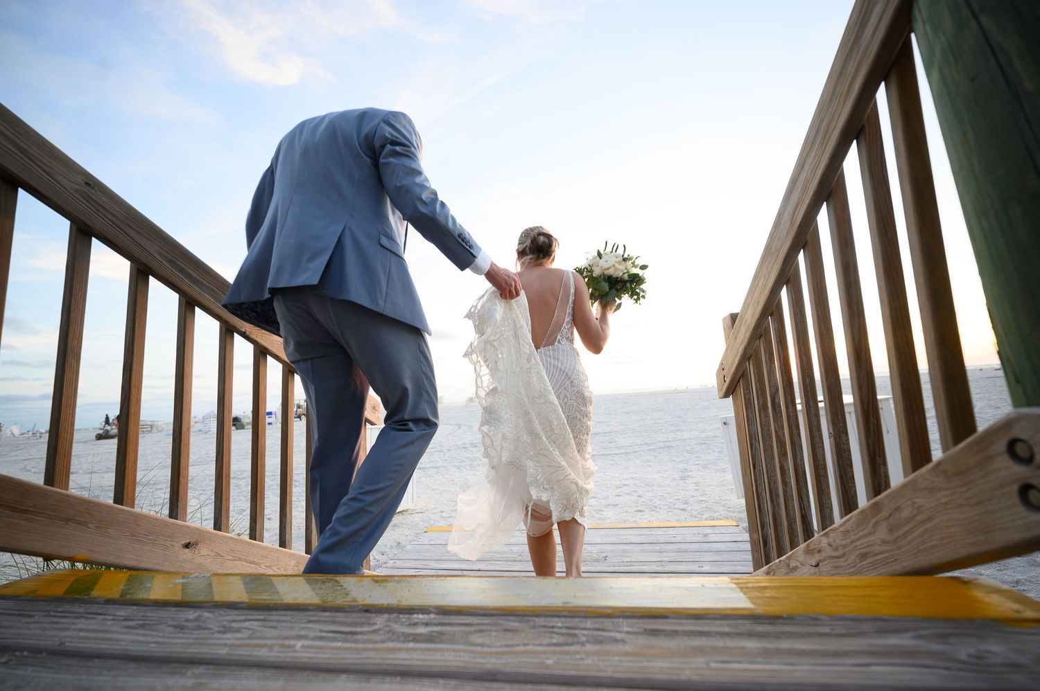 A couple descends wooden boardwalk stairs leading to a beach at their wedding celebration.