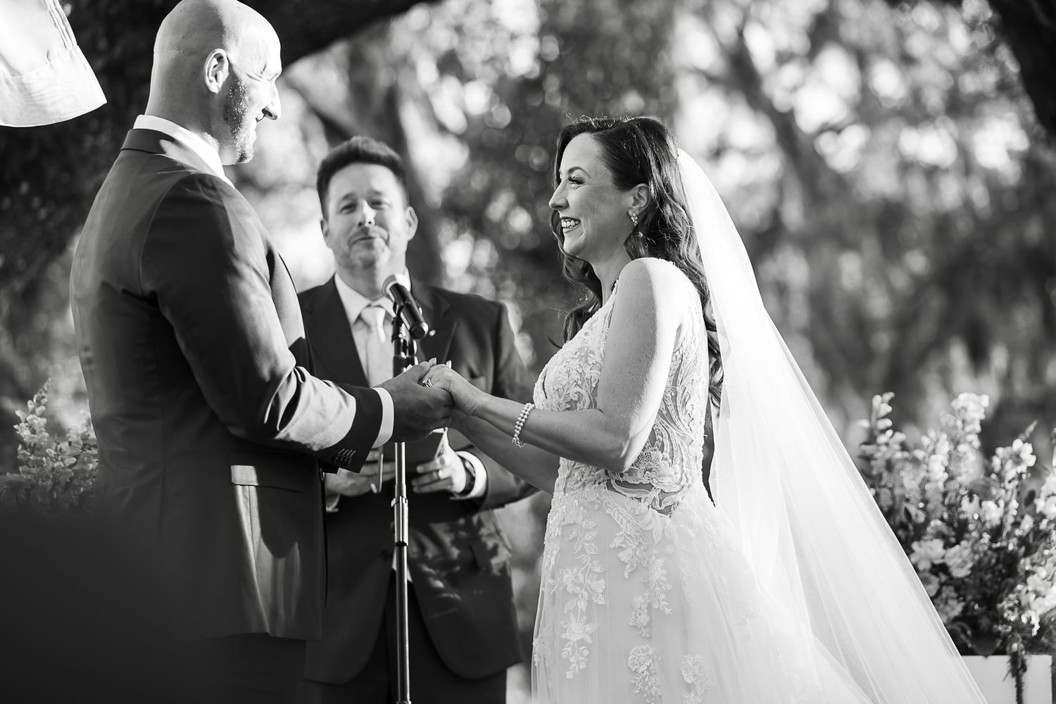 Black and white photo of a wedding ceremony with couple exchanging vows by an officiant outdoors.