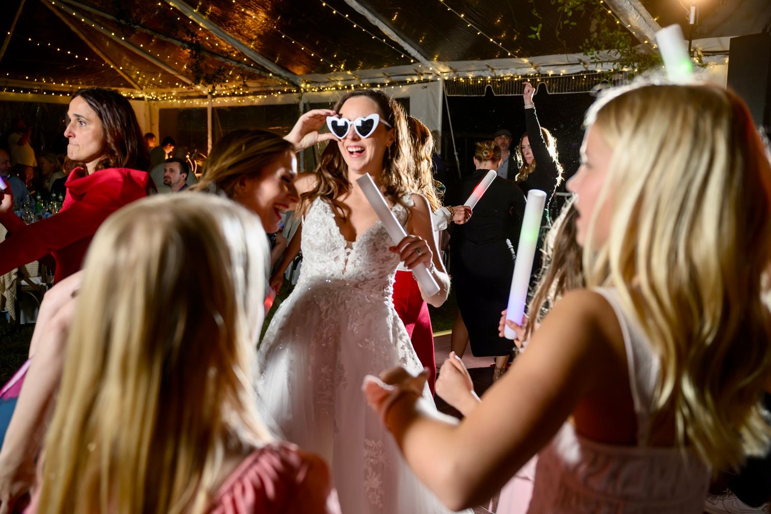 Wedding celebration on dance floor with guests holding glowing foam sticks under twinkle lights.