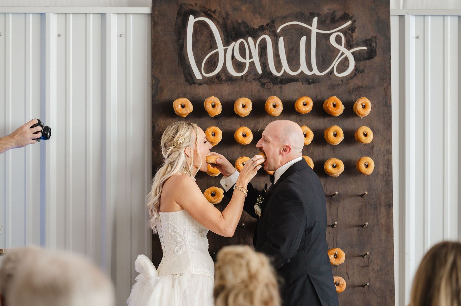 Bride and groom sharing a donut in front of a rustic wooden donut wall display at their wedding reception.