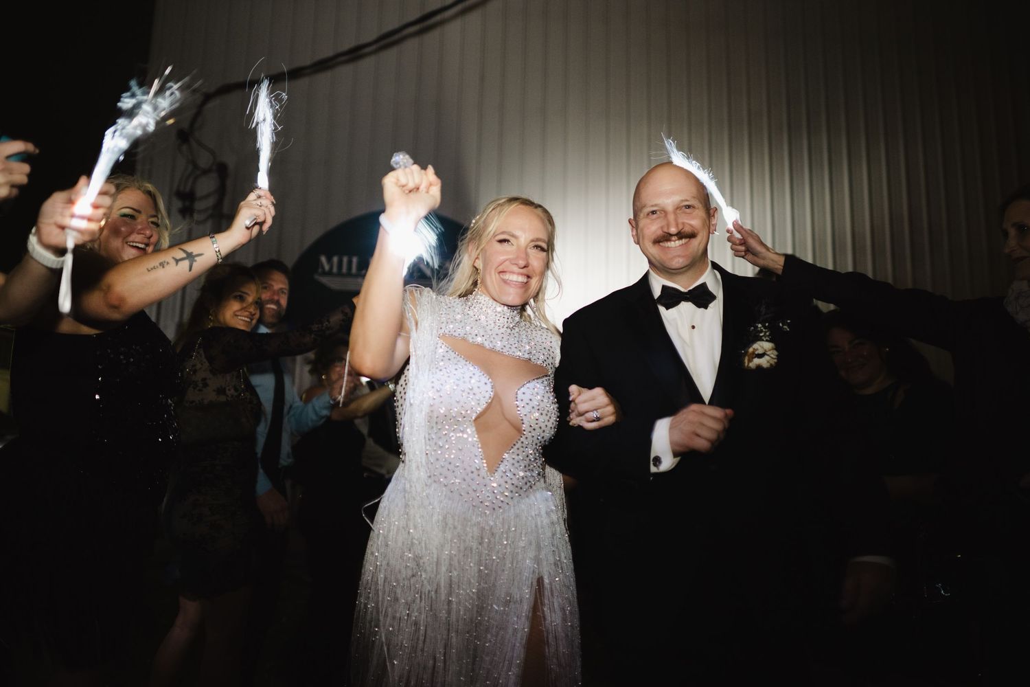 Bride and groom celebrating their wedding exit with sparklers surrounded by cheering guests at night.