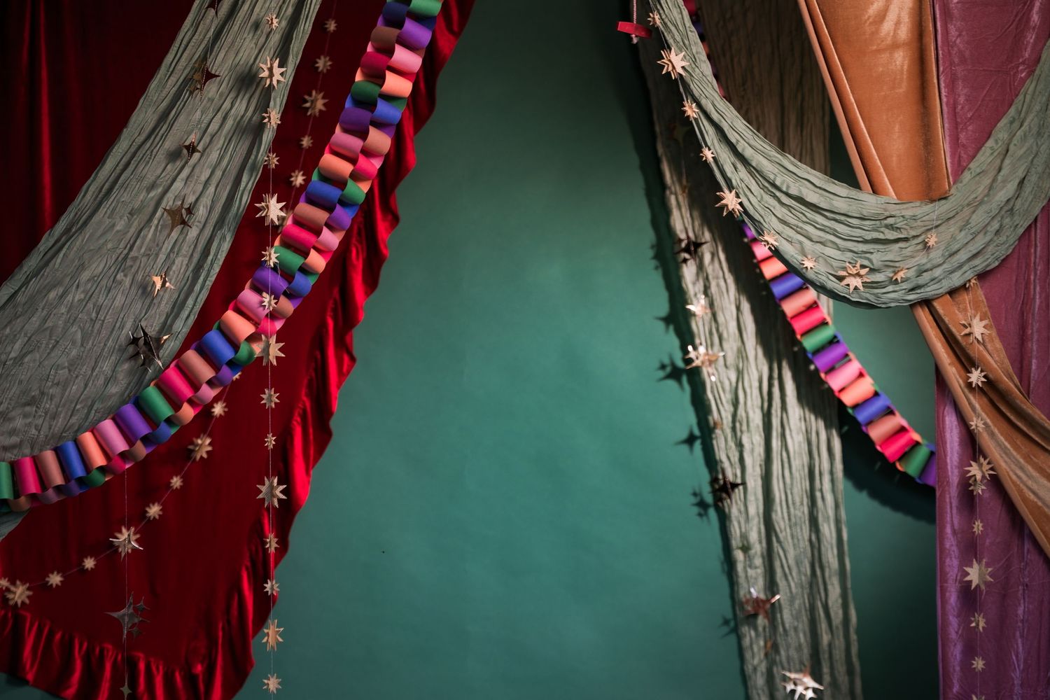 Colorful theatrical curtains with red velvet, gray fabric, multicolored tassels, and star embellishments against a teal wall.