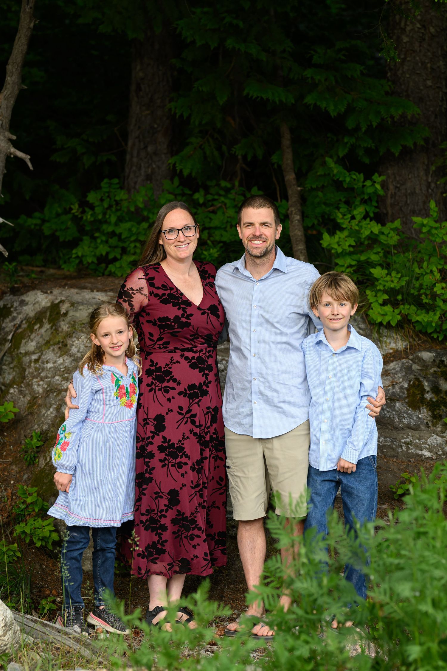 Family of four smiling outdoors in a forest setting, parents with two children posing for a portrait.