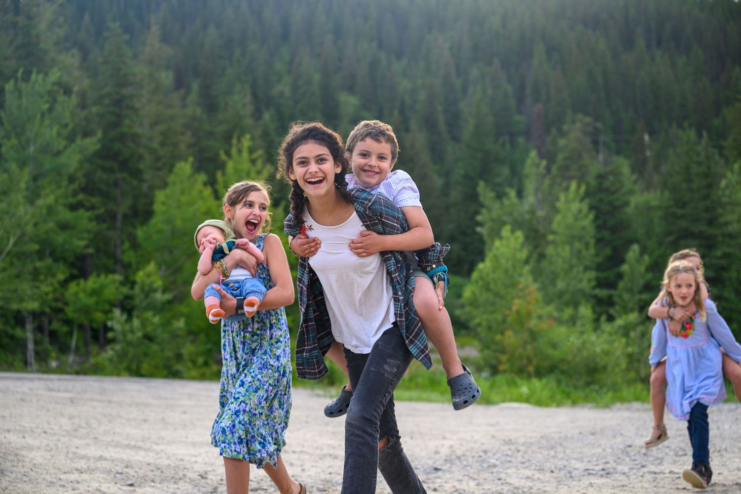 Joyful family with children piggyback riding outdoors near a lush green forest on a gravel path.