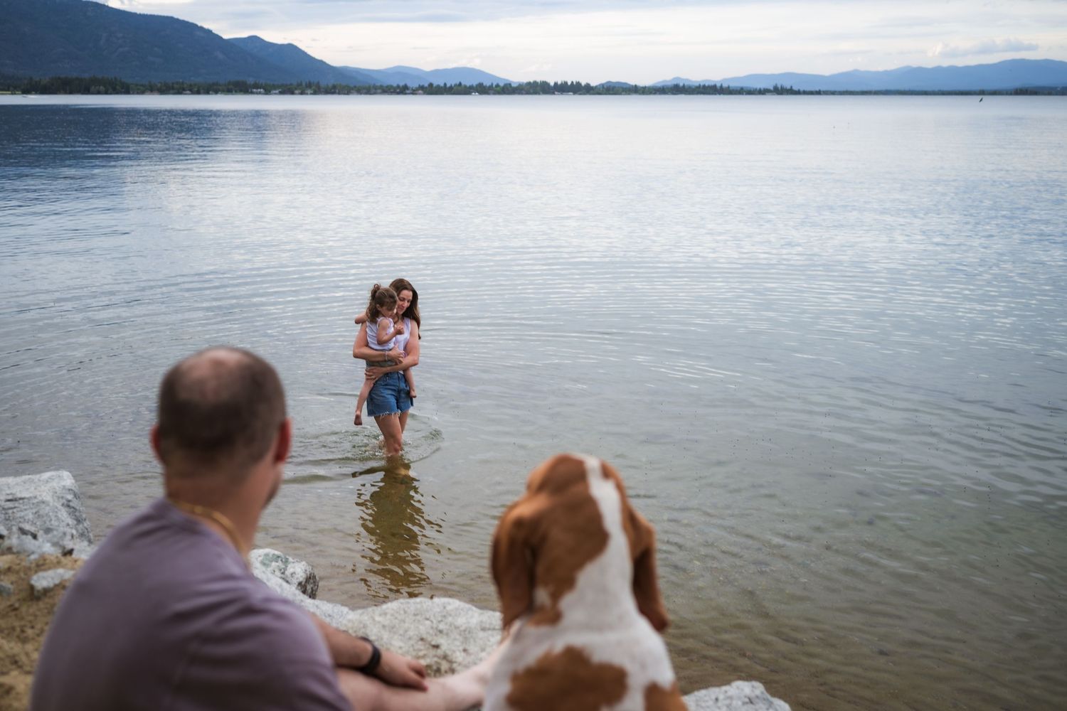 Man and dog watching woman carry child in shallow lake with mountain backdrop.