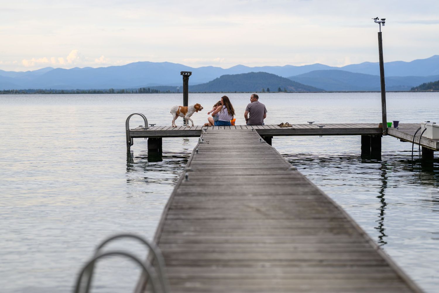Two people and a dog sit on a wooden dock overlooking a calm lake with mountain views in the background.