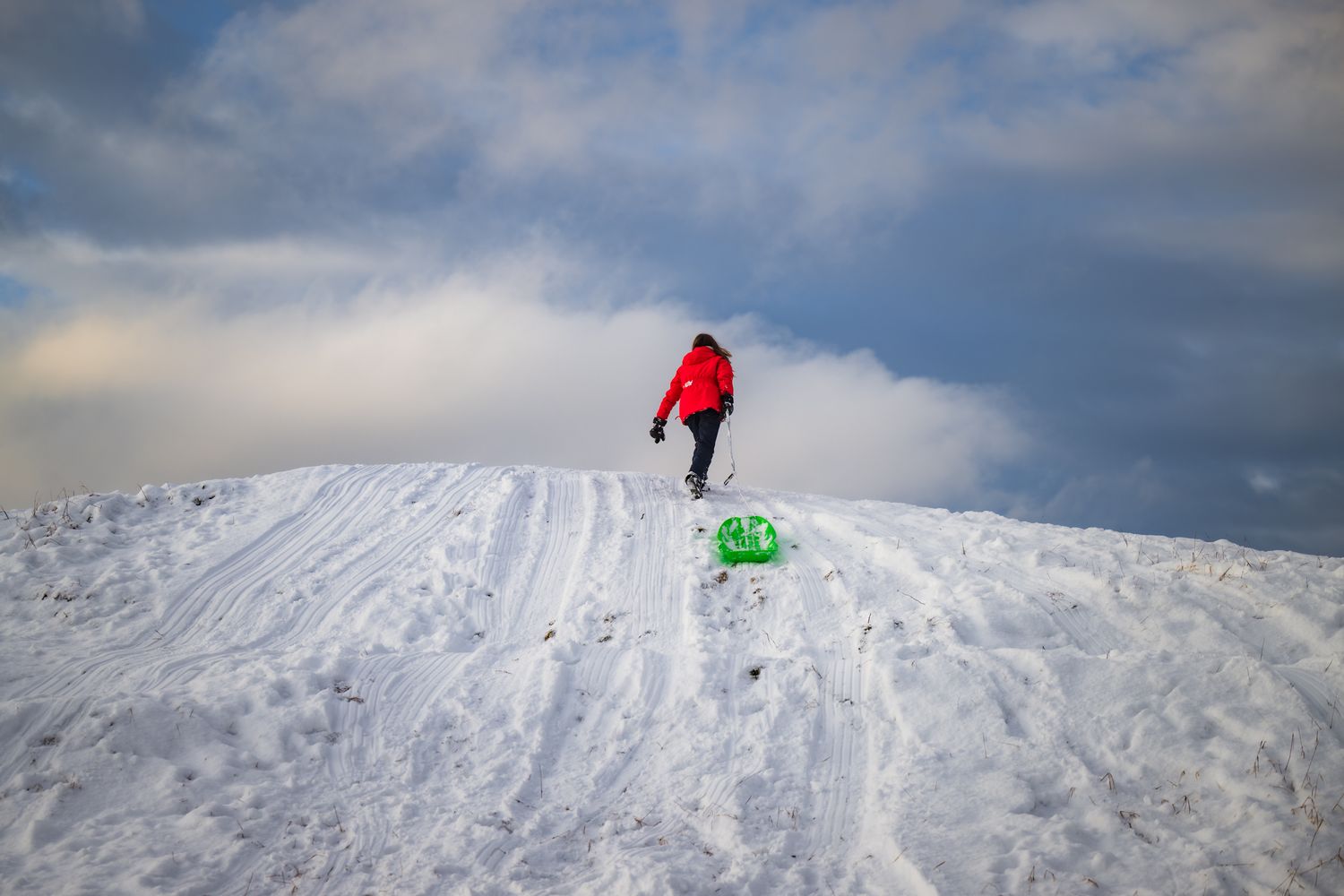 Girl in red jacket pulls a green sled up snowy slope against a cloudy mountain sky.